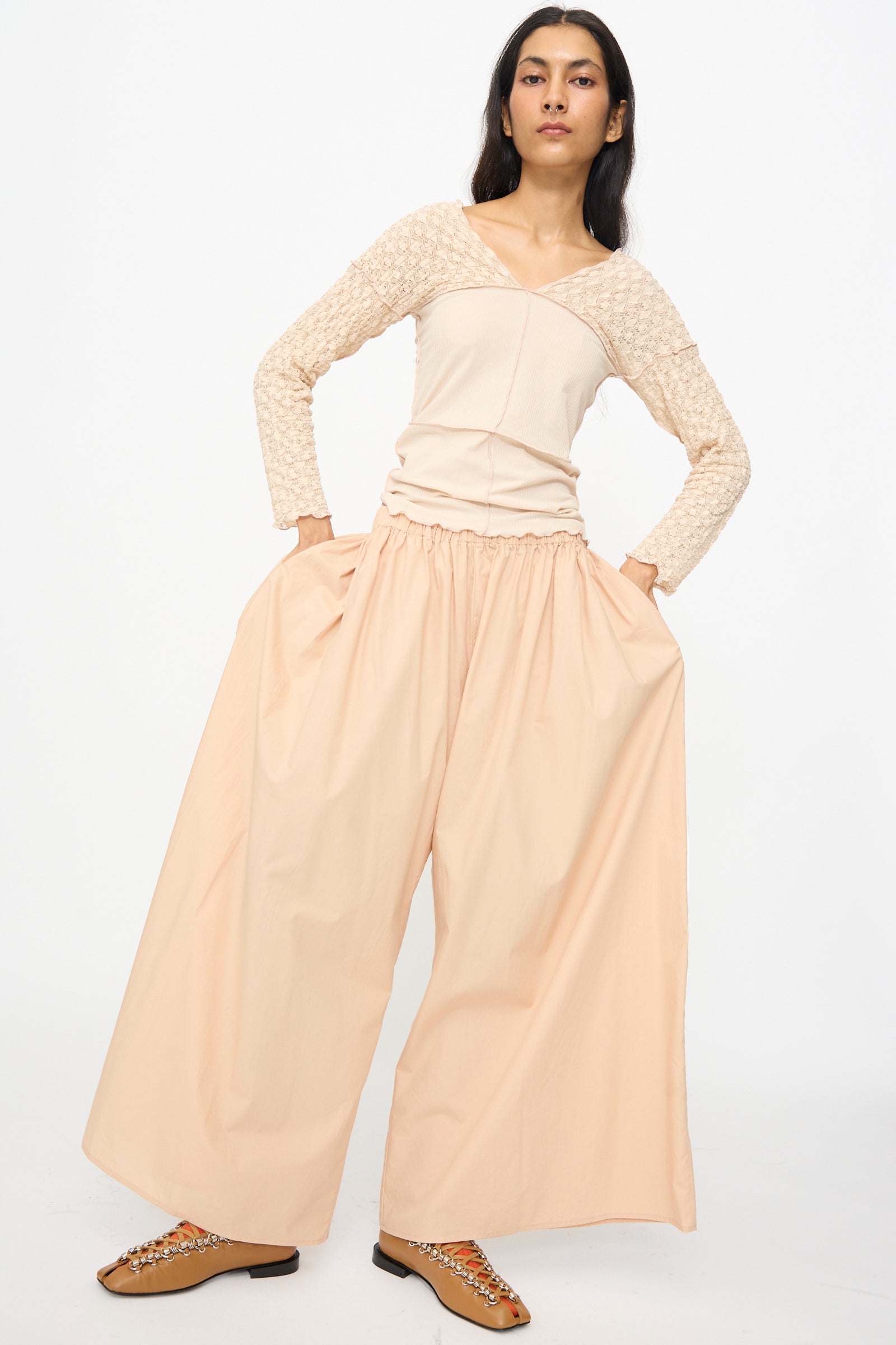 A person stands with hands in pockets, wearing Baserange Cable Pants in Andradita Sand—wide cotton pants with an elastic waist—paired with a textured long-sleeve top and embellished brown flats against a plain white background.