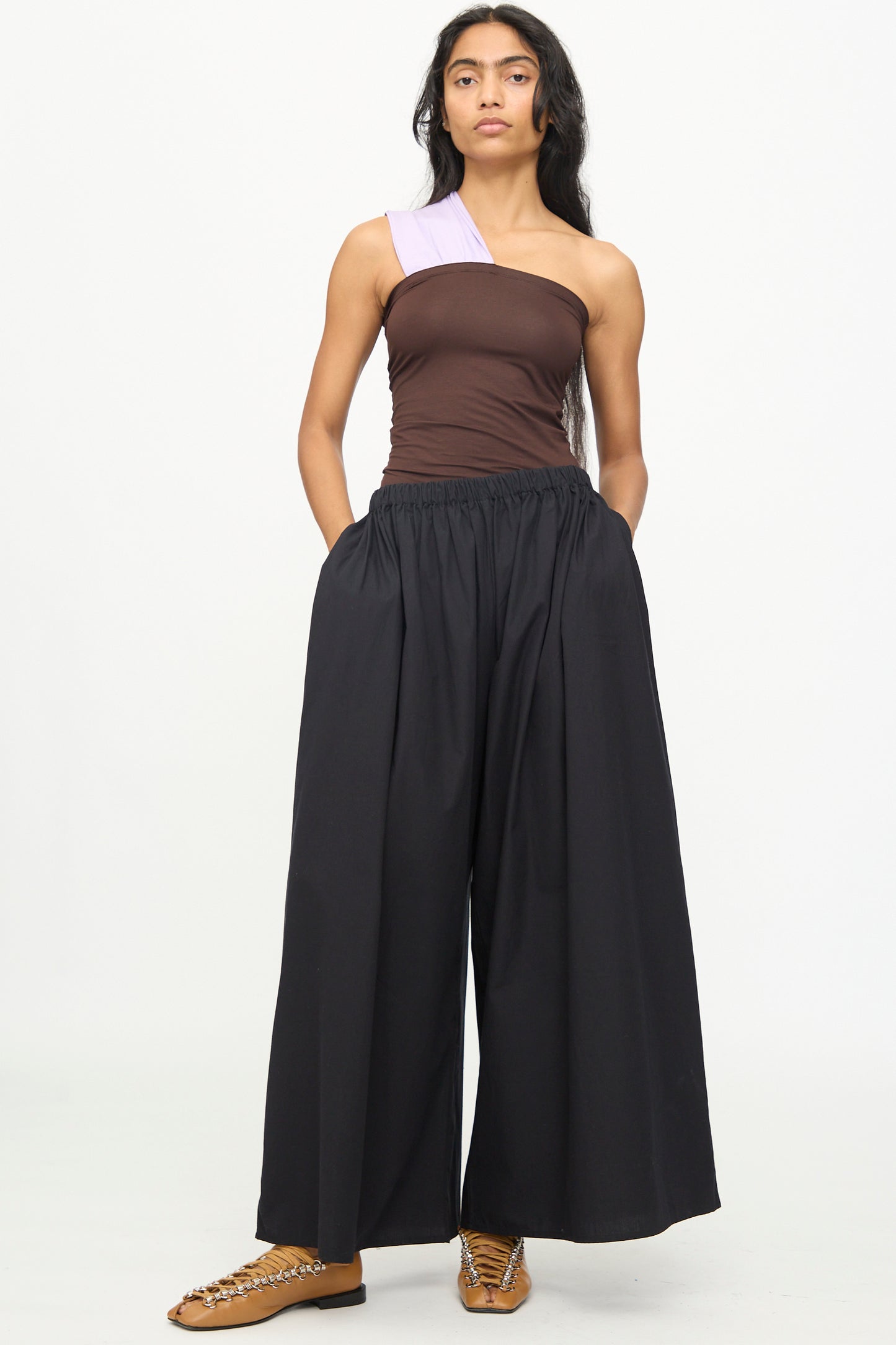 A woman stands against a white background wearing the Baserange Cable Pants in Black—wide cotton poplin pants with an elastic waist—paired with a brown one-shoulder top and tan shoes featuring a chain detail.