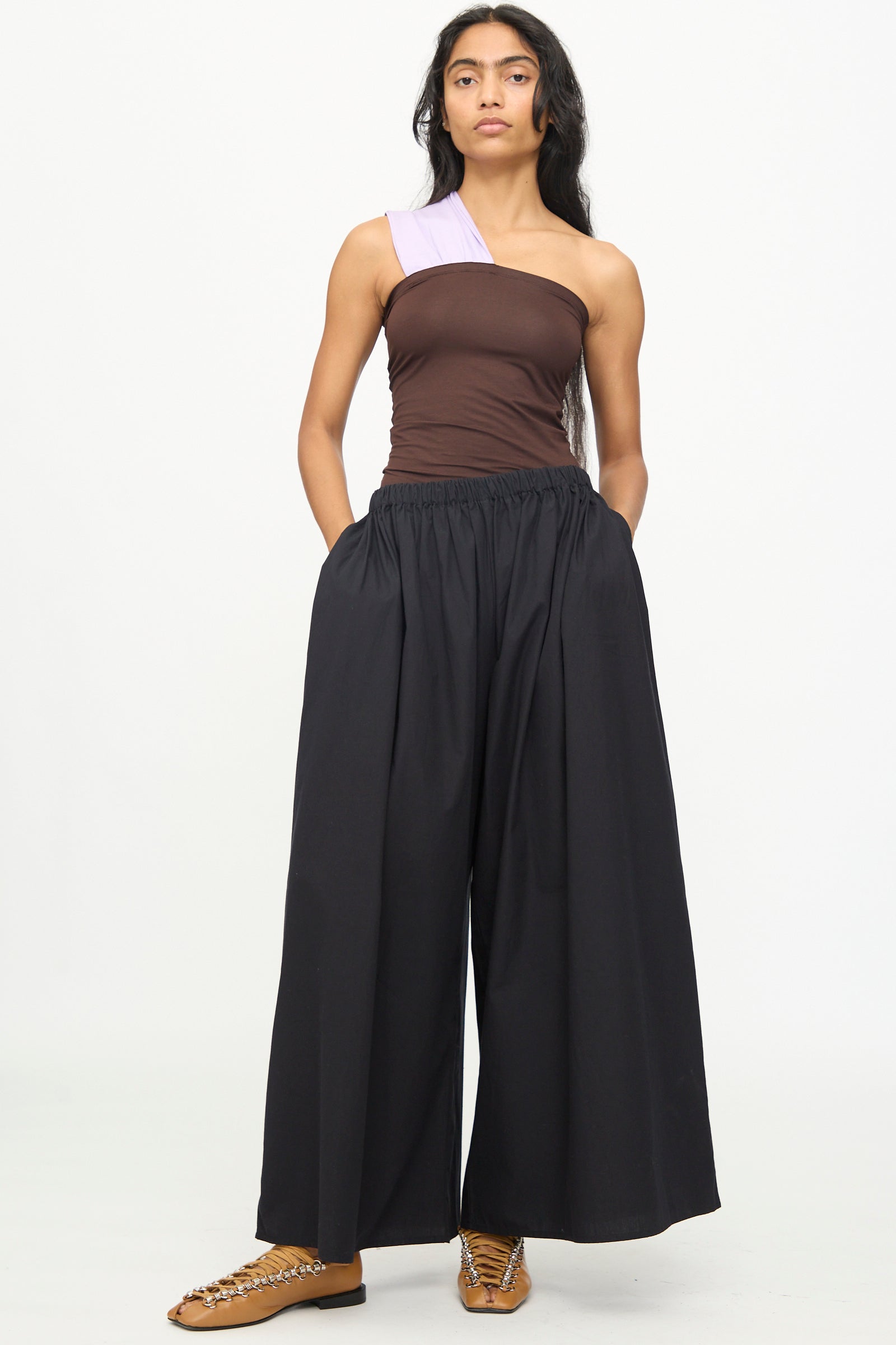 A woman stands against a white background wearing the Baserange Cable Pants in Black—wide cotton poplin pants with an elastic waist—paired with a brown one-shoulder top and tan shoes featuring a chain detail.