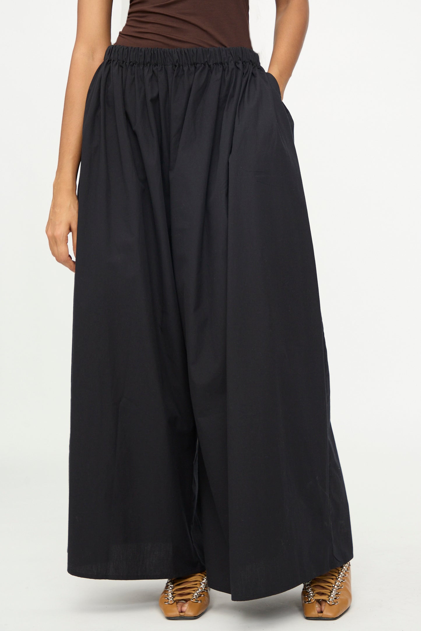 A person is wearing the Baserange Cable Pants in Black—poplin wide-leg pants with an elastic waist—paired with brown sandals, standing against a plain white background. The upper body is partially visible.