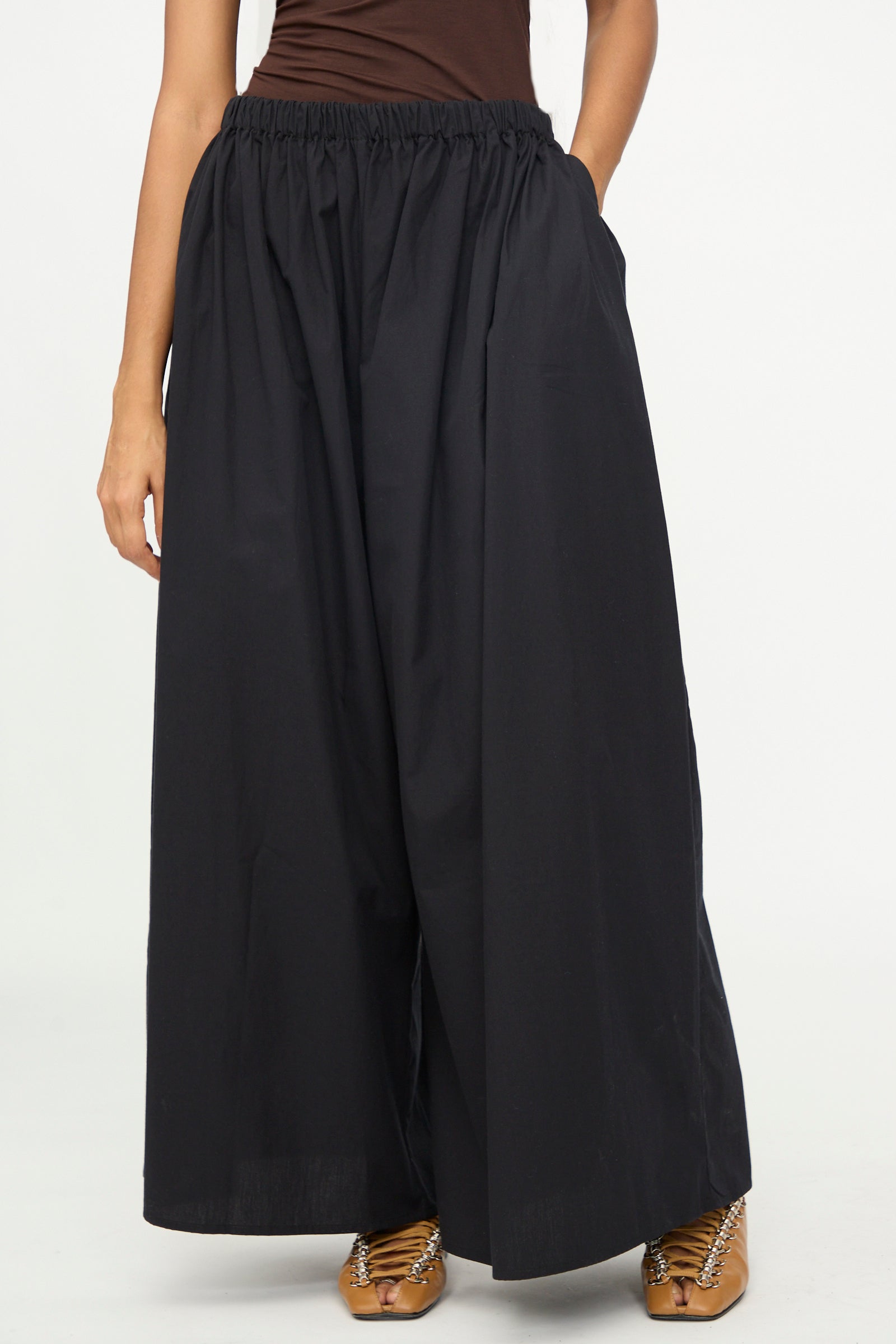 A person is wearing the Baserange Cable Pants in Black—poplin wide-leg pants with an elastic waist—paired with brown sandals, standing against a plain white background. The upper body is partially visible.