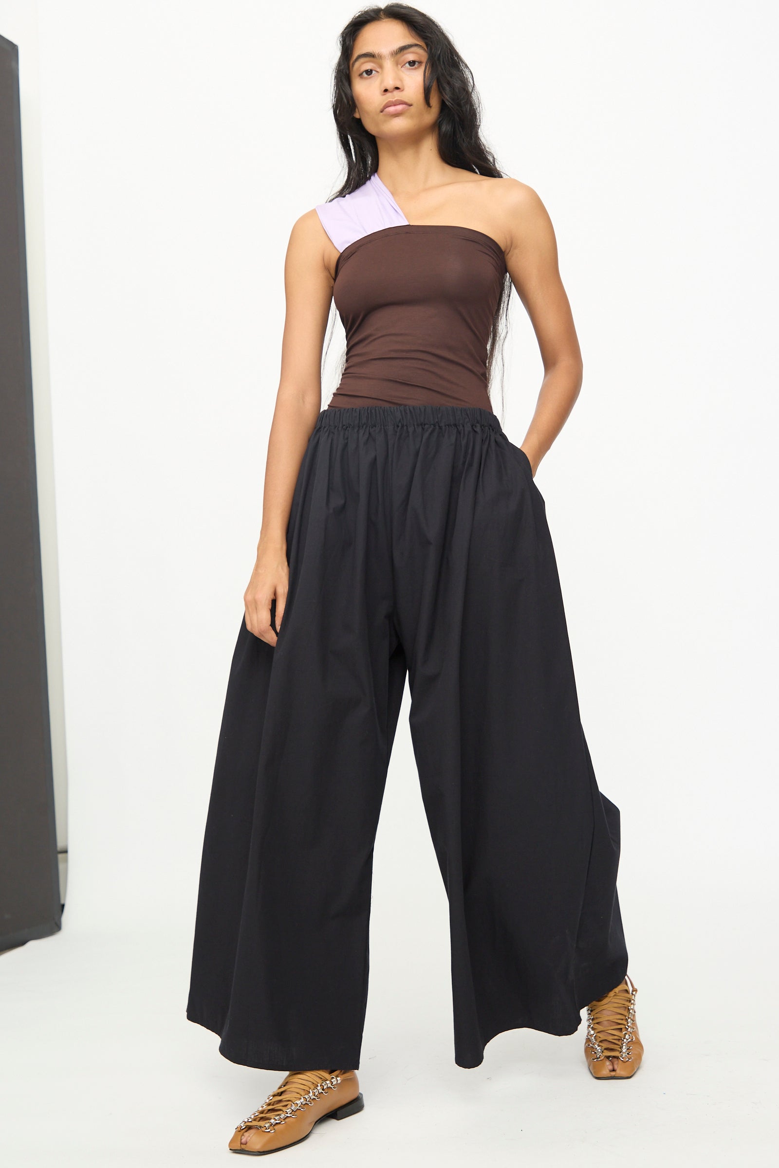 A person wearing a brown and lilac one-shoulder top pairs it with Baserange's Cable Pants in Black, which feature an elastic waist, and tan lace-up shoes while standing against a plain white background.