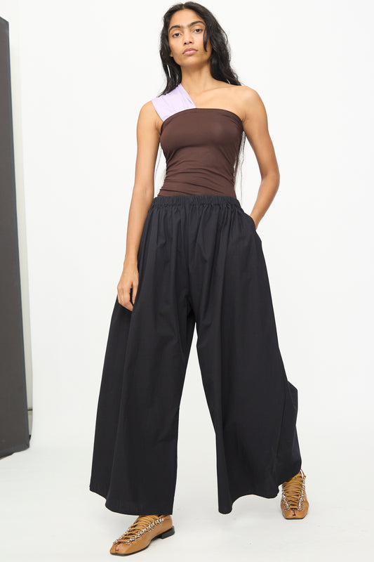A person wearing a brown and lilac one-shoulder top pairs it with Baserange's Cable Pants in Black, which feature an elastic waist, and tan lace-up shoes while standing against a plain white background.