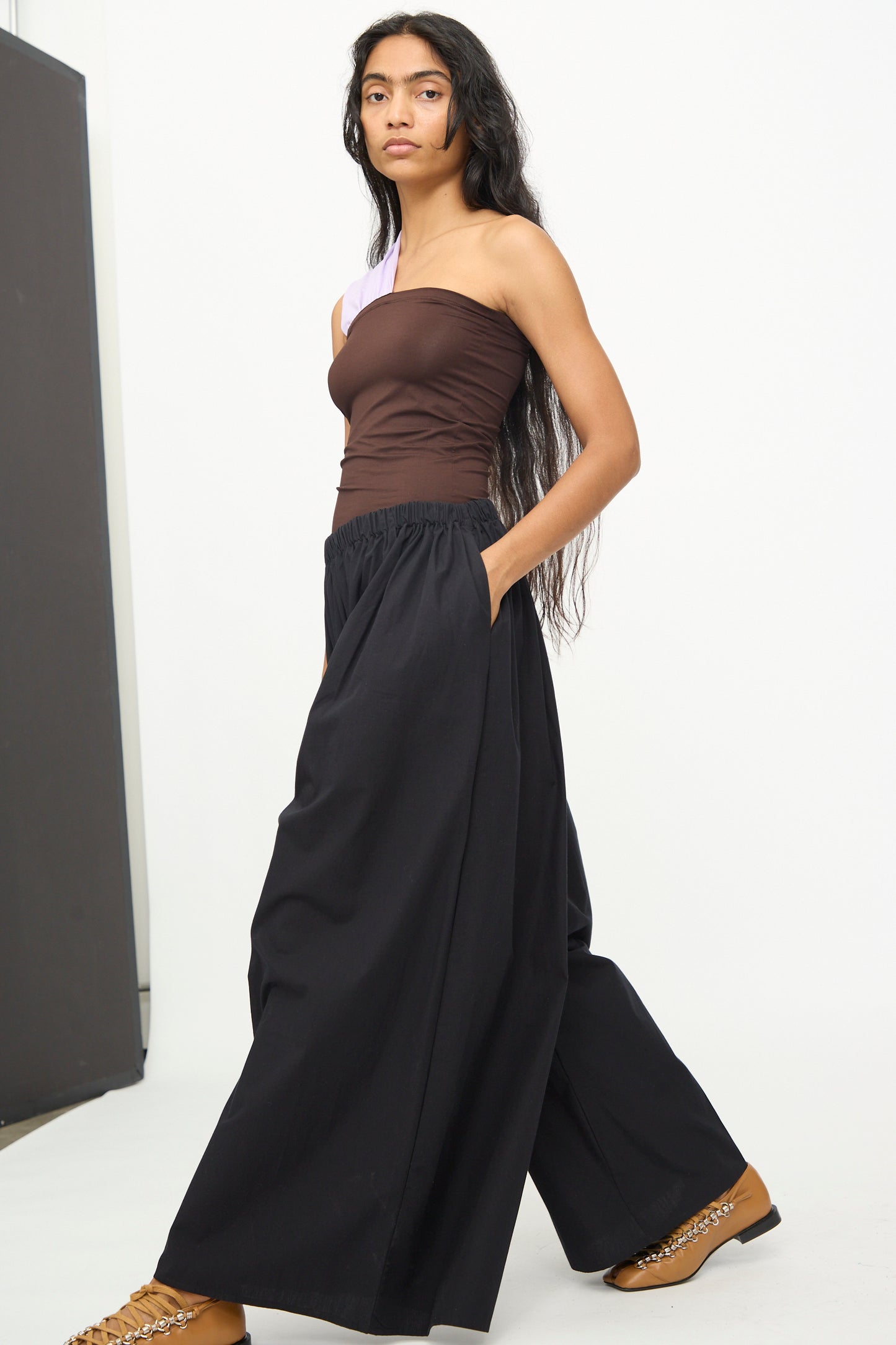 A person is wearing a brown one-shoulder top, Baserange Cable Pants in Black with an elastic waist, and tan shoes with chain detailing, standing against a plain white background.