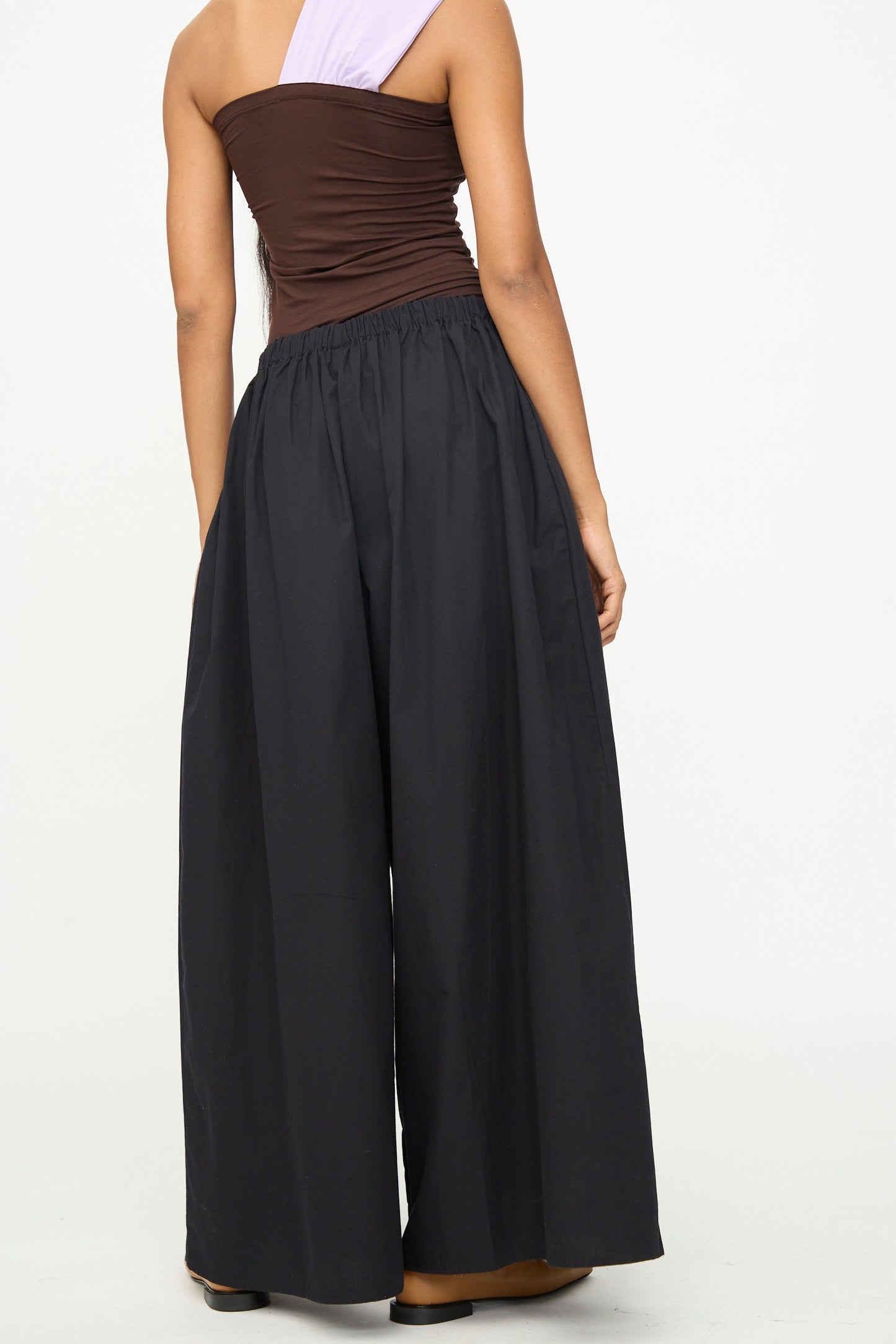 A person is seen from the back wearing Baserange’s Cable Pants in Black—high-waisted, wide-leg cotton poplin pants with an elastic waist—styled with a sleeveless brown and lavender top and standing against a plain white background.