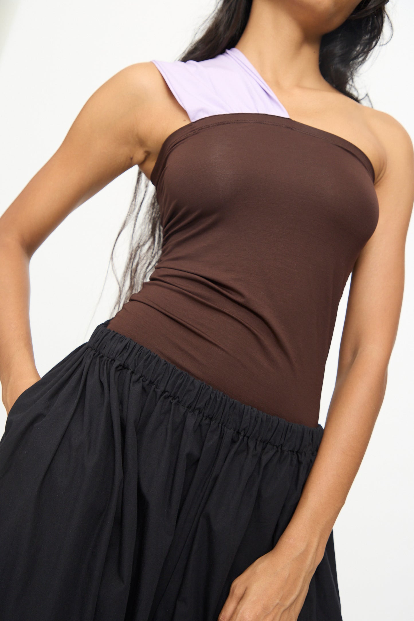 A woman stands against a plain background with one hand in her pocket, wearing a brown one-shoulder top with a lavender strap and Baserange's Cable Pants in Black, featuring wide cotton poplin legs and an elastic waist.