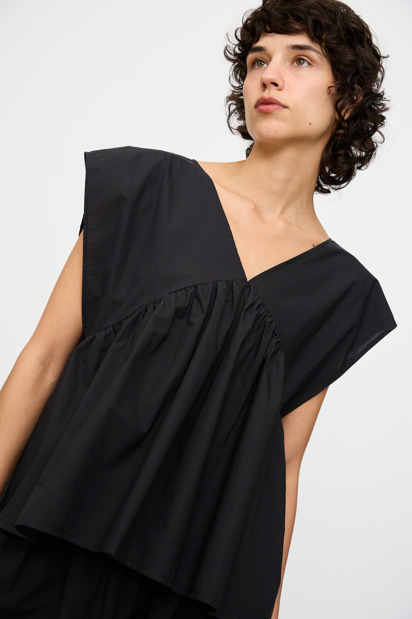 Someone wears the Baserange Cable Top in Black, an oversized, sleeveless organic cotton piece with a deep V-neck and gathered details, standing against a plain light background.
