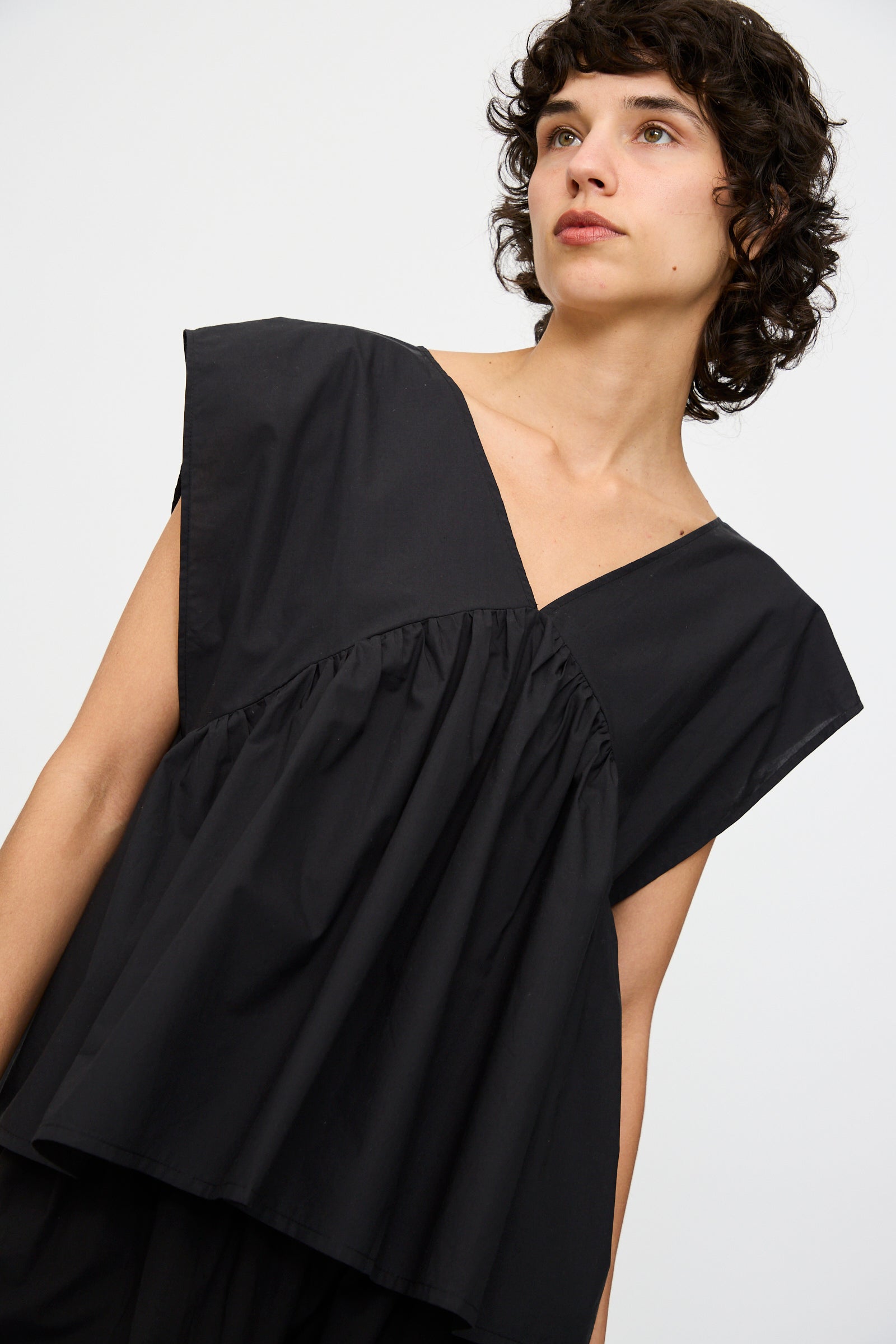Someone wears the Baserange Cable Top in Black, an oversized, sleeveless organic cotton piece with a deep V-neck and gathered details, standing against a plain light background.