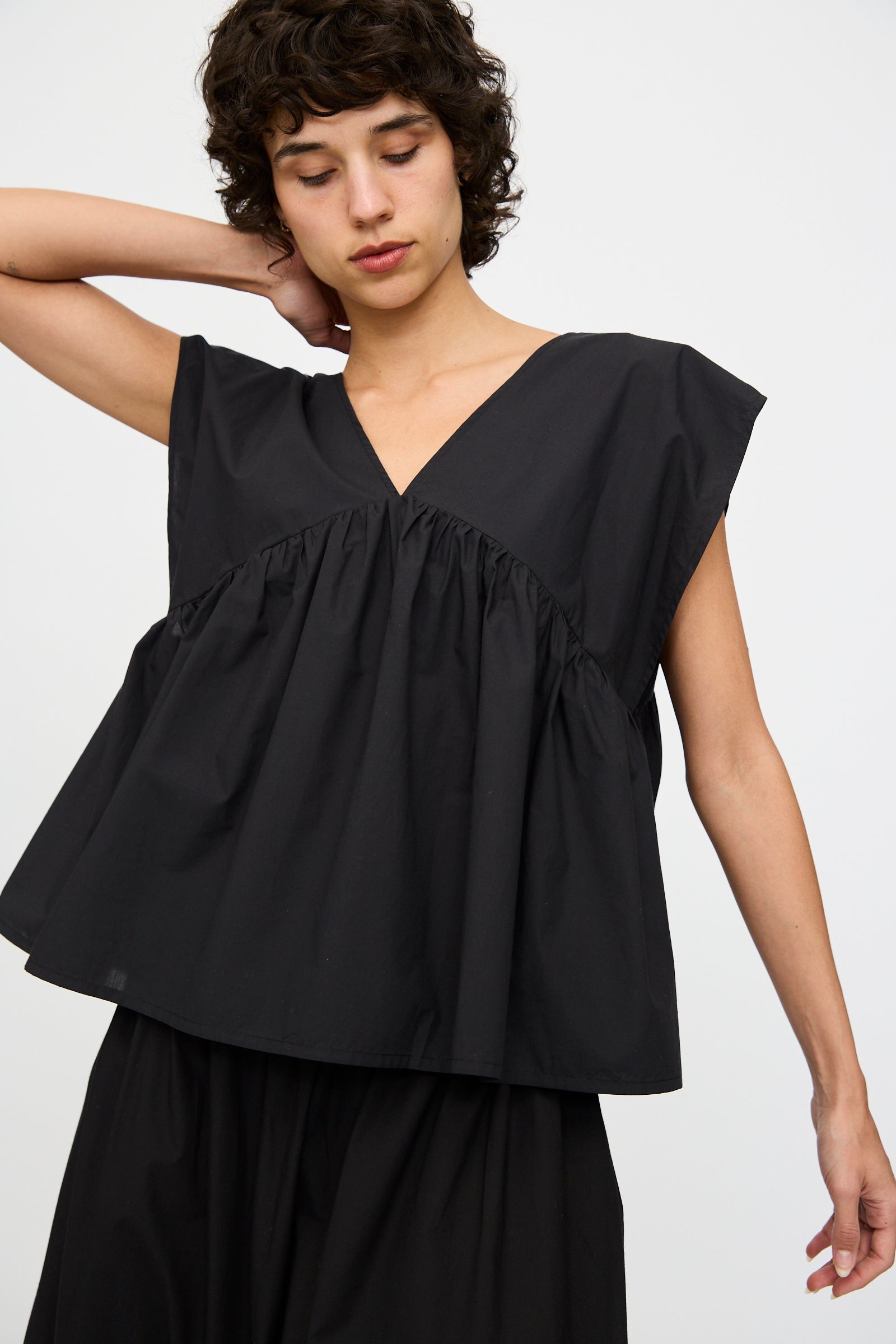 A person stands against a plain background, wearing Baserange's Cable Top in Black—an oversized fit with wide sleeves and a gathered waist—paired with a matching black skirt made from organic cotton.