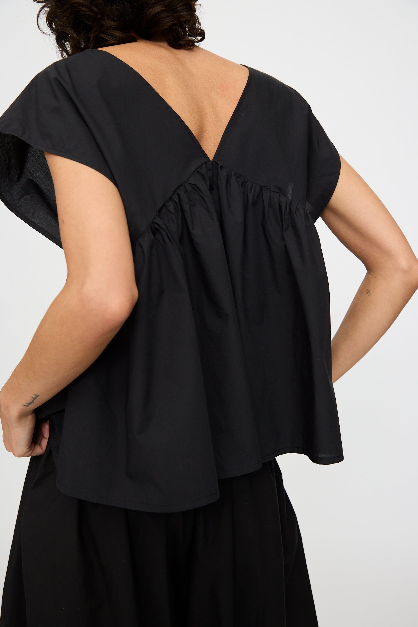 A person shown from behind wears the Baserange Cable Top in Black—an oversized organic cotton short-sleeve with a V-shaped back.