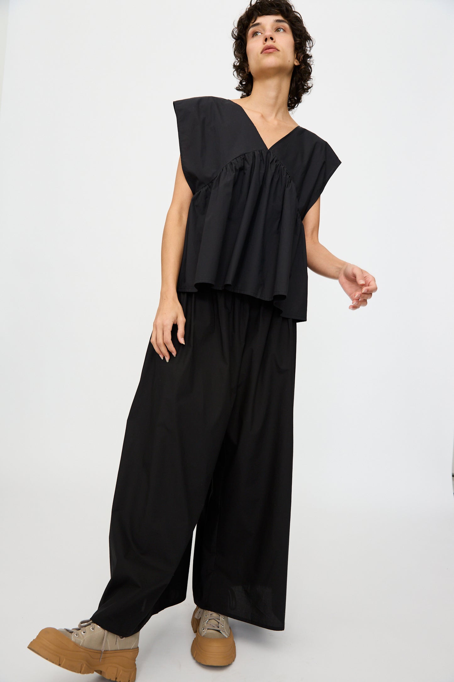 A person wears the Baserange Cable Top in Black, styled oversized and sleeveless, with wide-leg black pants and tan platform shoes, standing against a plain white background.