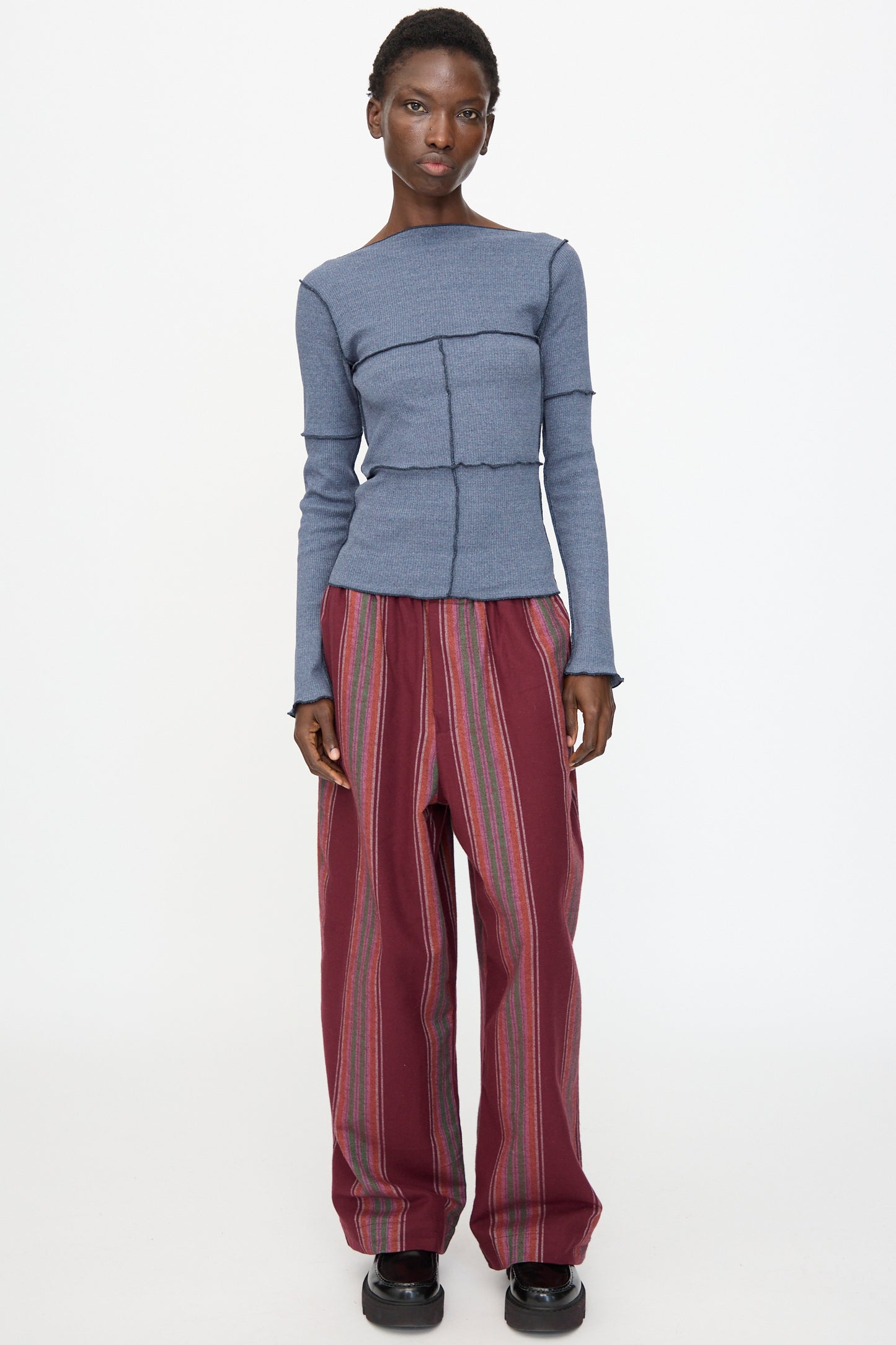 A person stands against a plain background, wearing the Baserange Cinder Long Sleeve Tee in Dark Denim paired with loose maroon striped pants and black shoes.