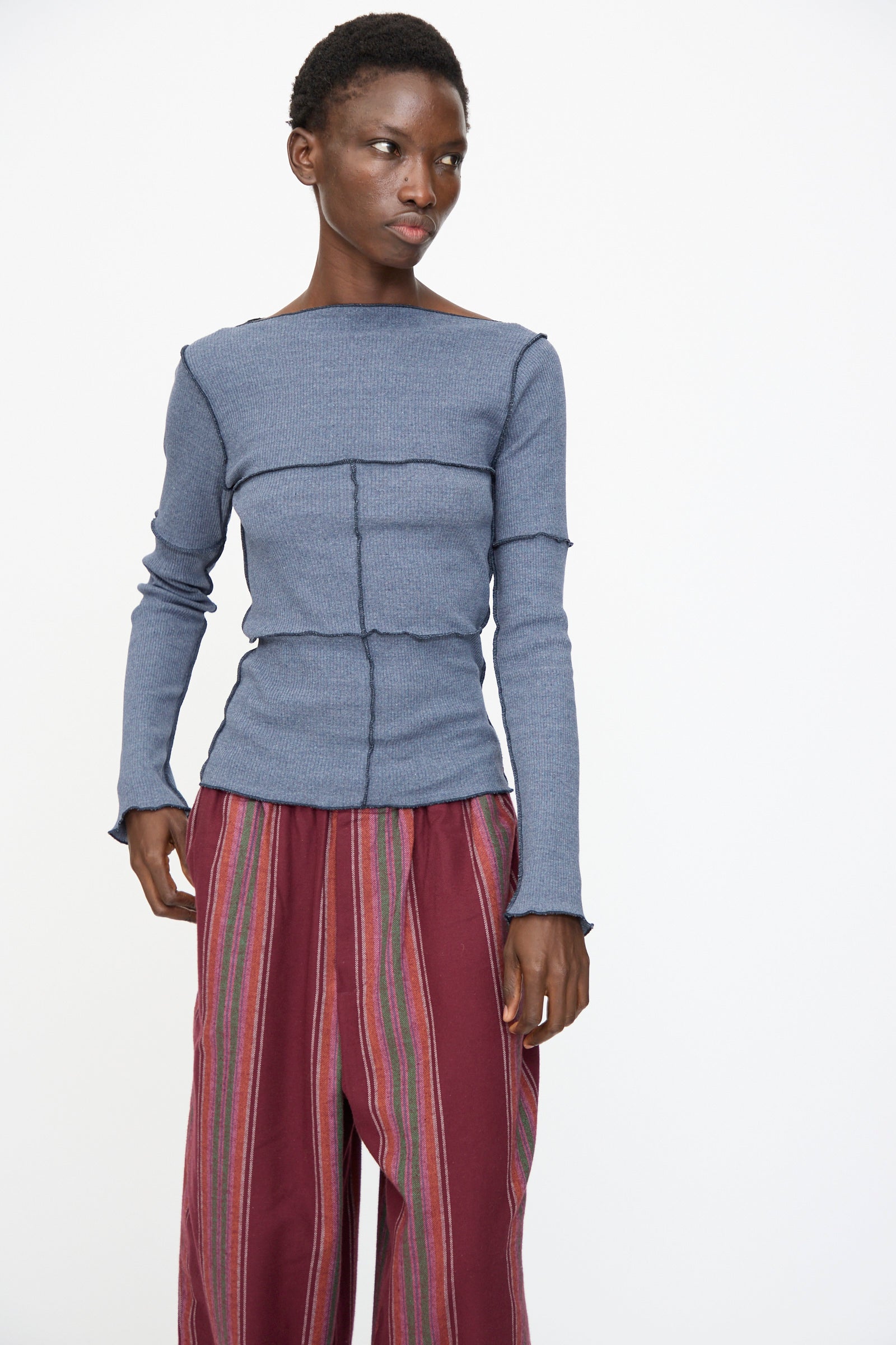 Model wears the Baserange Cinder Long Sleeve Tee in Dark Denim with loose maroon striped pants, standing against a plain white background.