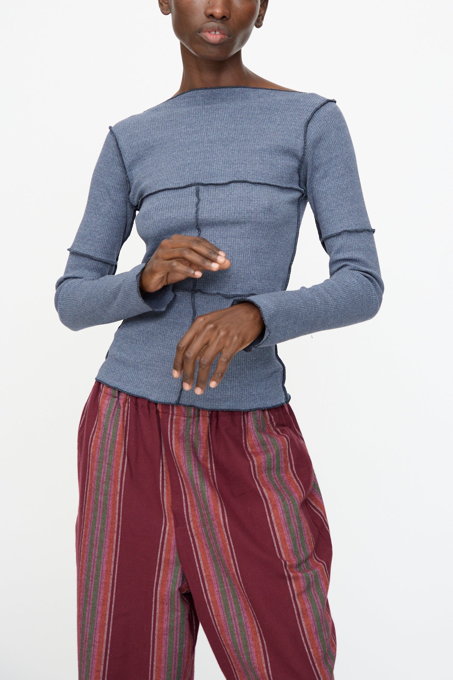 A person wearing the Baserange Cinder Long Sleeve Tee in Dark Denim, styled with high-waisted maroon vertical striped pants, stands against a plain white background.