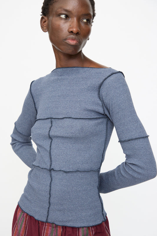 A person wears the Baserange Cinder Long Sleeve Tee in Dark Denim, a slim fit organic cotton top with exposed seams, paired with maroon patterned pants, standing against a plain white background.