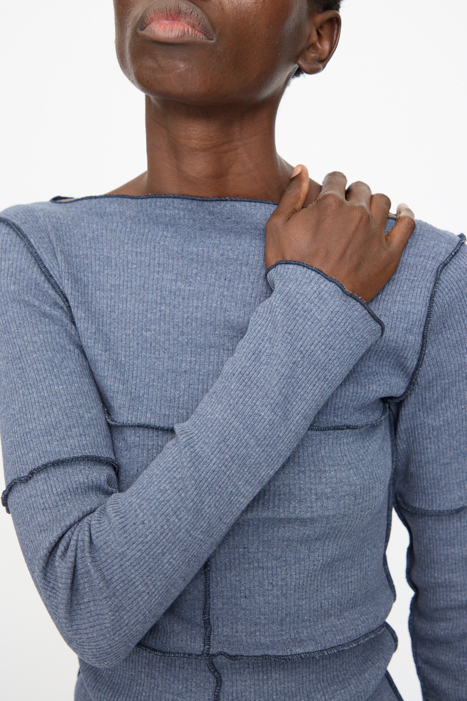 A person wears the Baserange Cinder Long Sleeve Tee in Dark Denim, a blue slim fit top with ribbed texture and visible seams, touching their shoulder. Only the lower half of their face is visible.