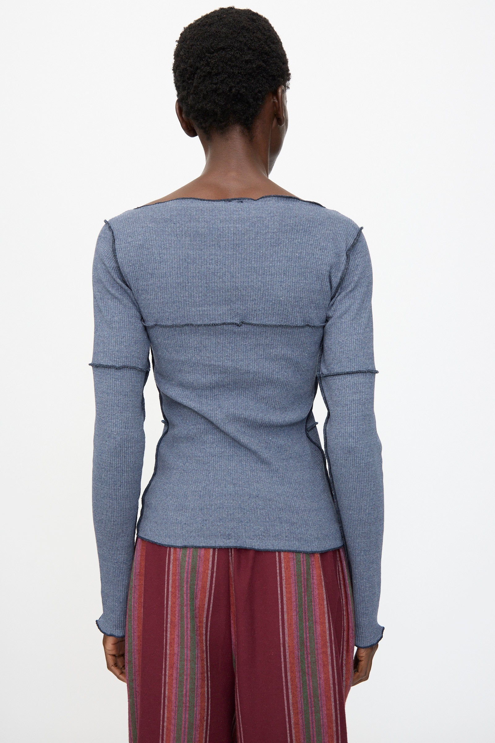 A person stands facing away, wearing the Baserange Cinder Long Sleeve Tee in Dark Denim, a slim fit blue ribbed top with visible seams, paired with red striped pants against a plain white background.