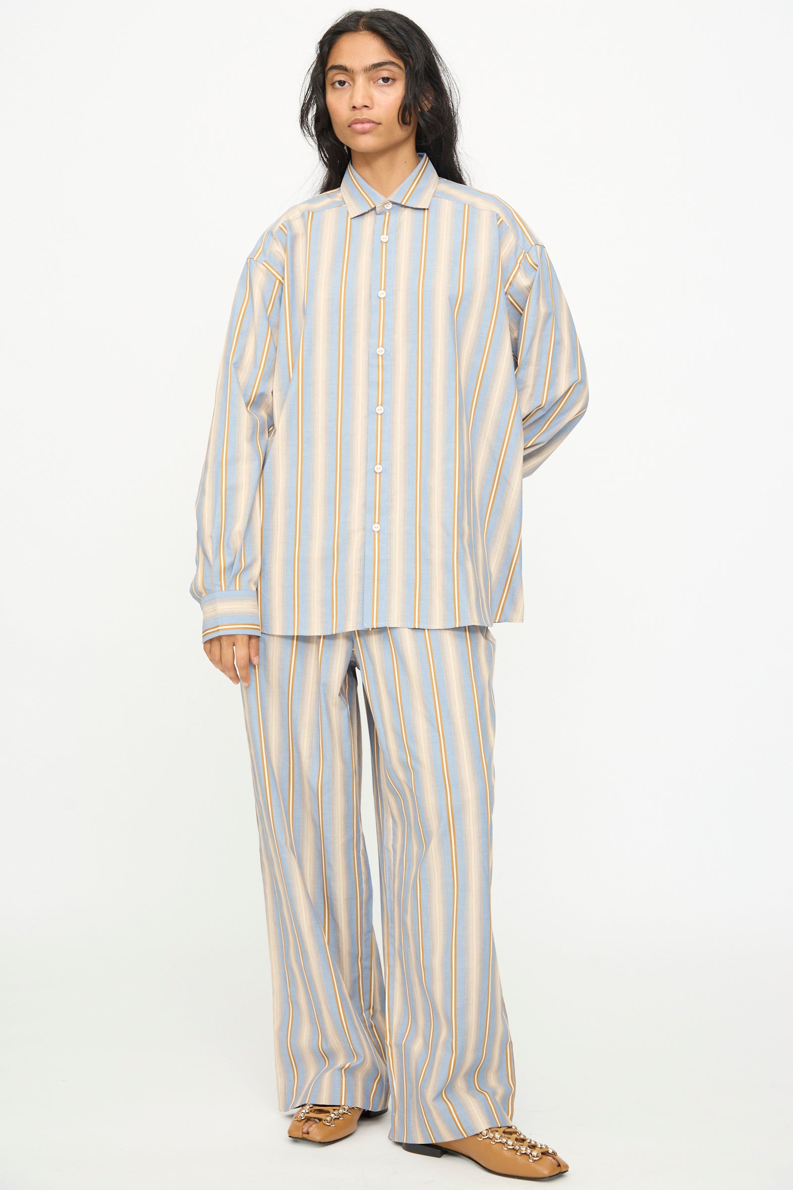 A person stands against a plain background, wearing the Baserange Claude Pants in Grid Stripe—a pair of loose-fitting, wide leg pants with vertical blue, yellow, and white stripes—paired with a matching long-sleeve, button-up shirt and brown shoes.