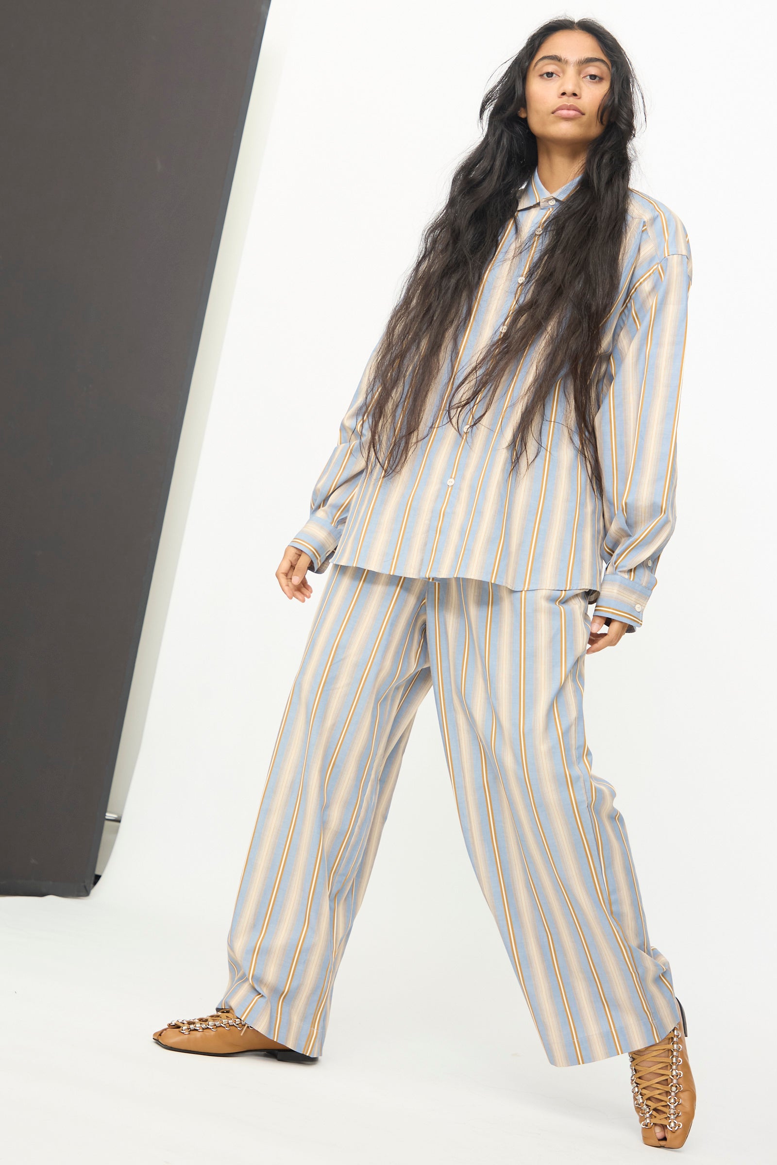 Person standing against a white background, wearing the Baserange Claude Pants in Grid Stripe—loose-fitting, striped wide leg pants—paired with a matching button-up shirt and tan lace-up shoes.