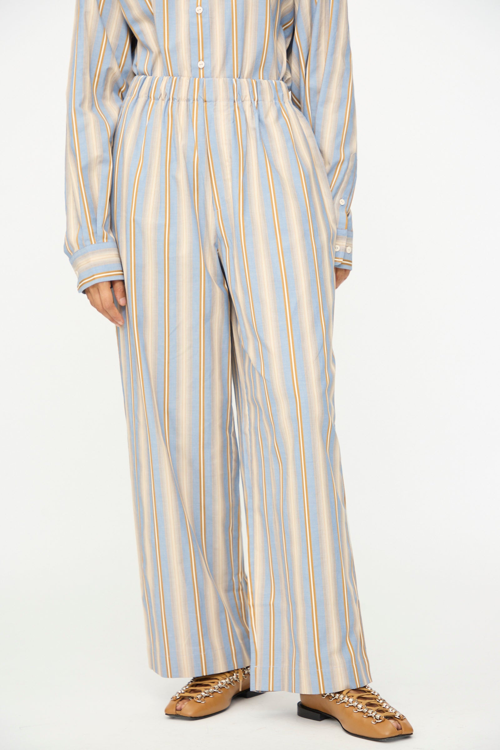 A person, shown from the shoulders down against a plain background, wears the Baserange Claude Pants in Grid Stripe—loose, high-waisted pull-on pants made from light blue and yellow vertical striped organic cotton—paired with a matching shirt and tan shoes featuring chain details.