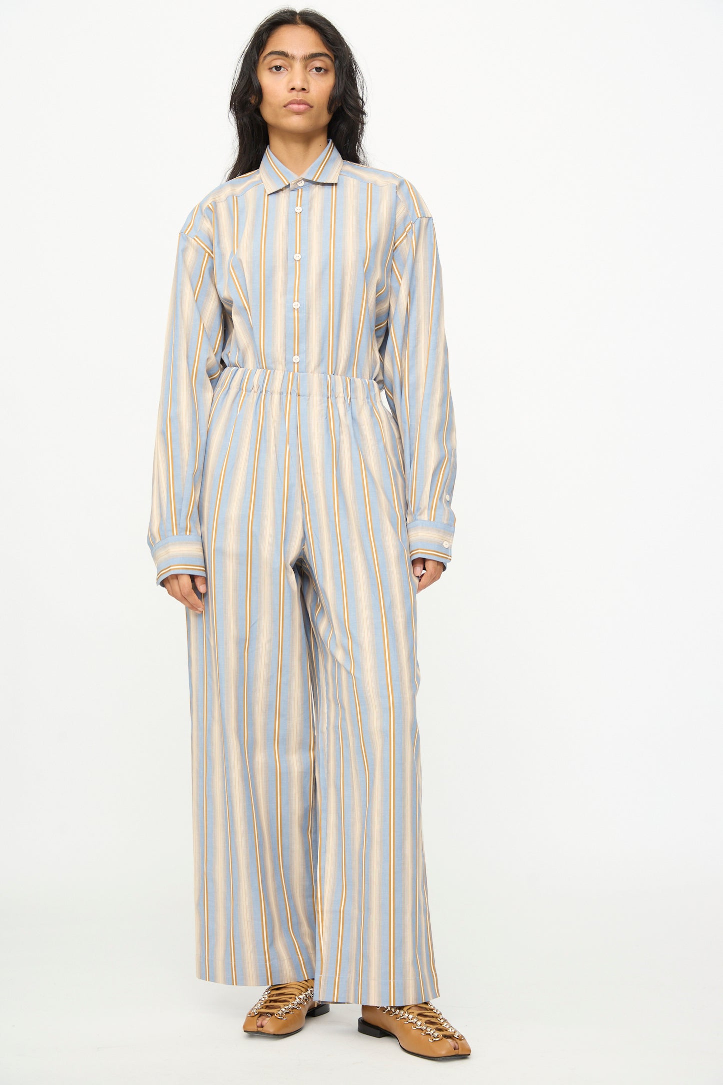 A person stands against a plain background, wearing Baserange's Claude Pants in Grid Stripe—loose-fitting, organic cotton pull-on wide leg pants—paired with a matching striped long-sleeved shirt and brown shoes.