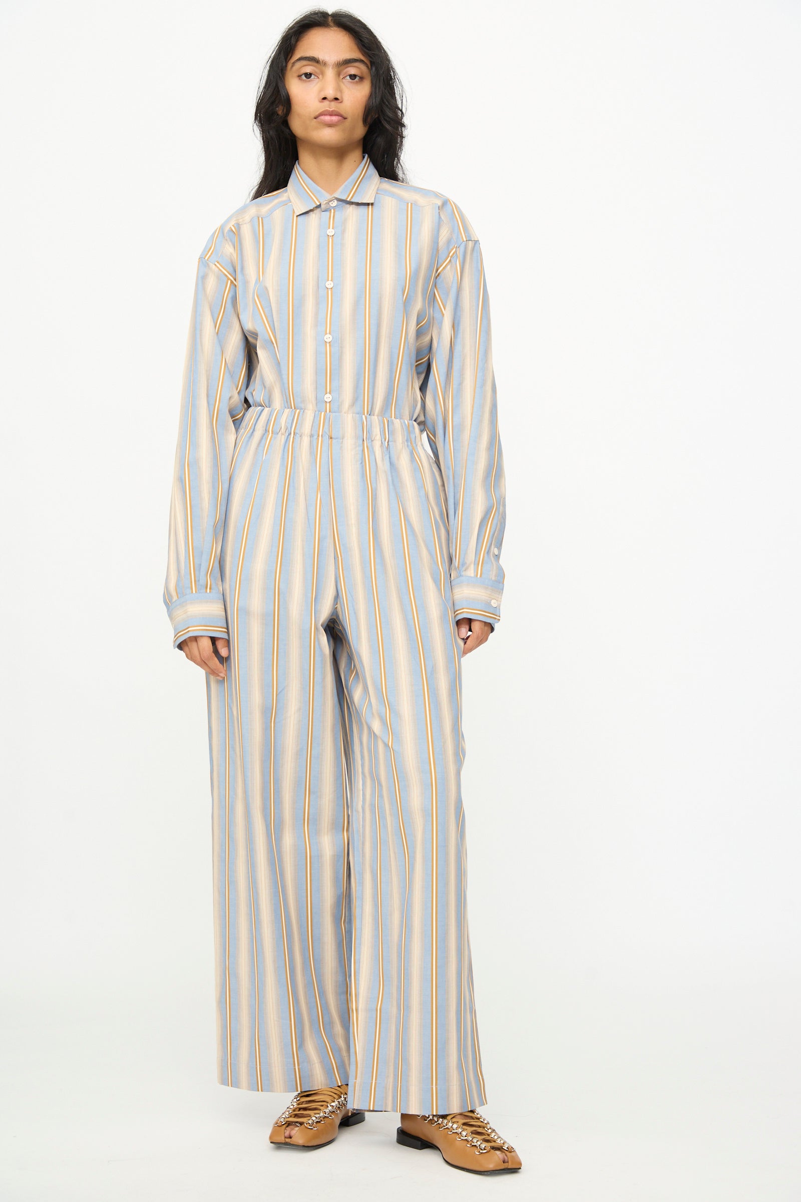 A person stands against a plain background, wearing Baserange's Claude Pants in Grid Stripe—loose-fitting, organic cotton pull-on wide leg pants—paired with a matching striped long-sleeved shirt and brown shoes.