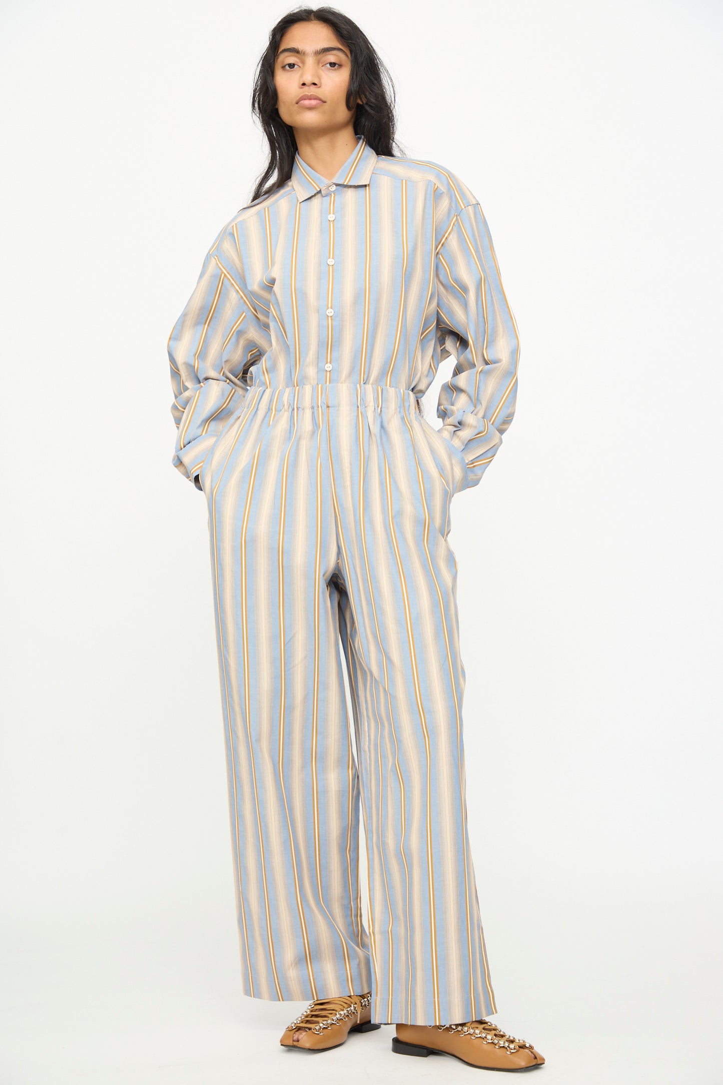 A person wearing the Baserange Claude Pants in Grid Stripe—a loose-fitting, wide-leg style featuring vertical blue, yellow, and white stripes—paired with tan loafers and standing with hands in pockets against a plain background.
