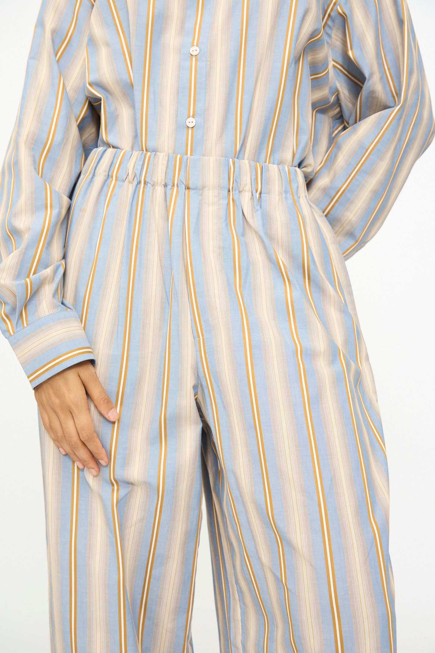 A person wearing Baserange’s Claude Pants in Grid Stripe—a pair of loose-fitting, wide leg pants with blue, beige, and yellow stripes—styled with a coordinating long-sleeve, button-up shirt.