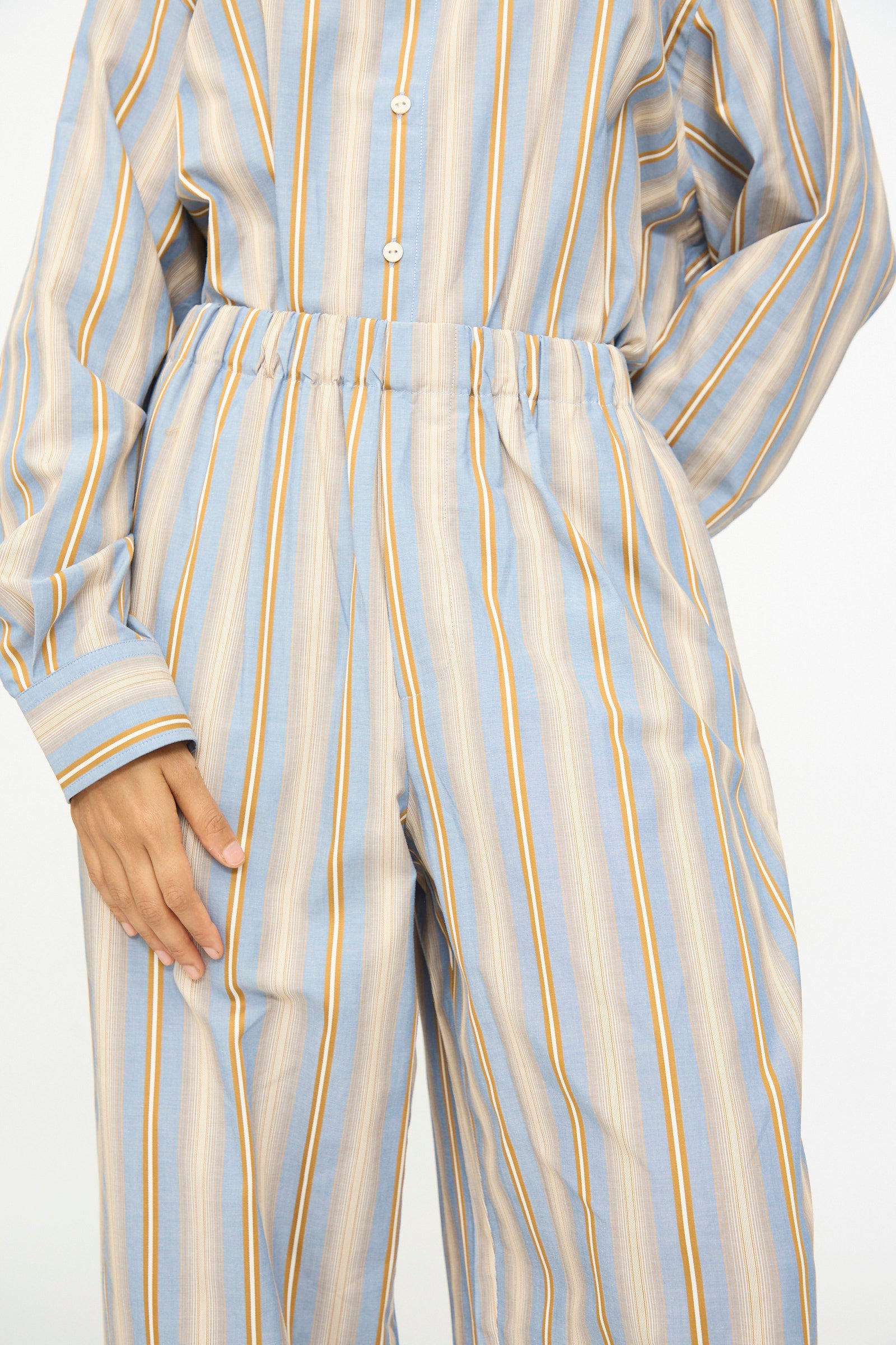 A person wearing Baserange’s Claude Pants in Grid Stripe—a pair of loose-fitting, wide leg pants with blue, beige, and yellow stripes—styled with a coordinating long-sleeve, button-up shirt.