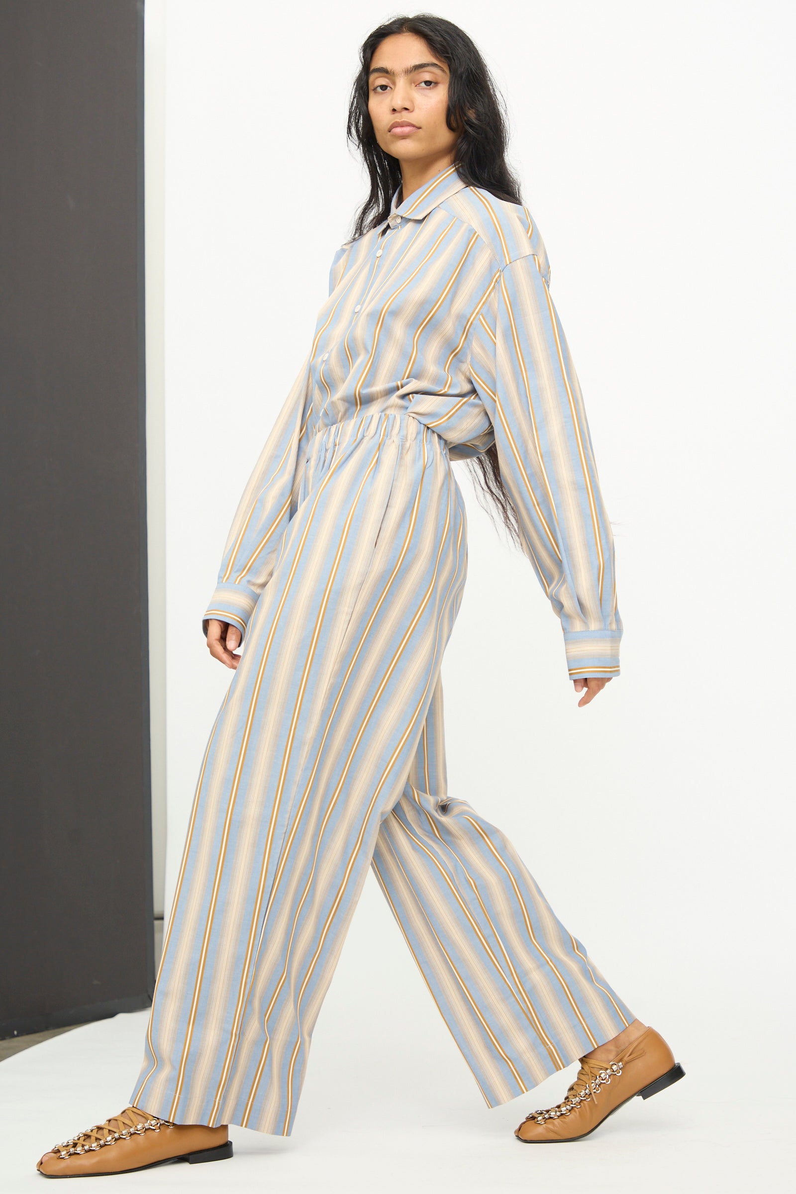 A person wearing the Baserange Claude Pants in Grid Stripe, styled with a matching loose, long-sleeved shirt and tan shoes, walks against a plain background.