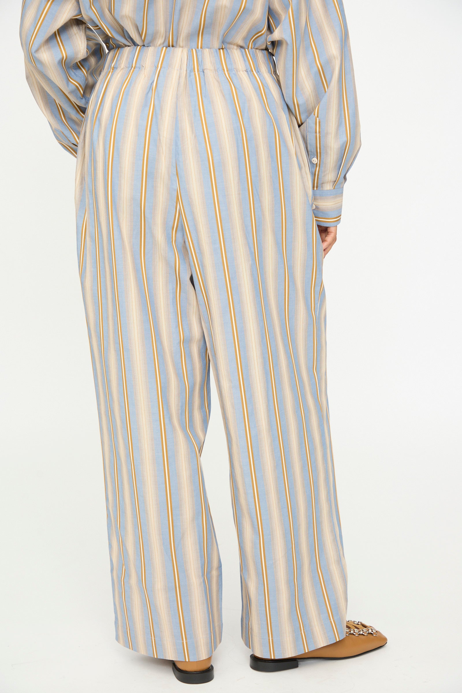 A person wearing Baserange Claude Pants in Grid Stripe—a loose-fitting, high-waisted pull-on style with vertical yellow and blue stripes—viewed from the back; brown shoes visible at the bottom.