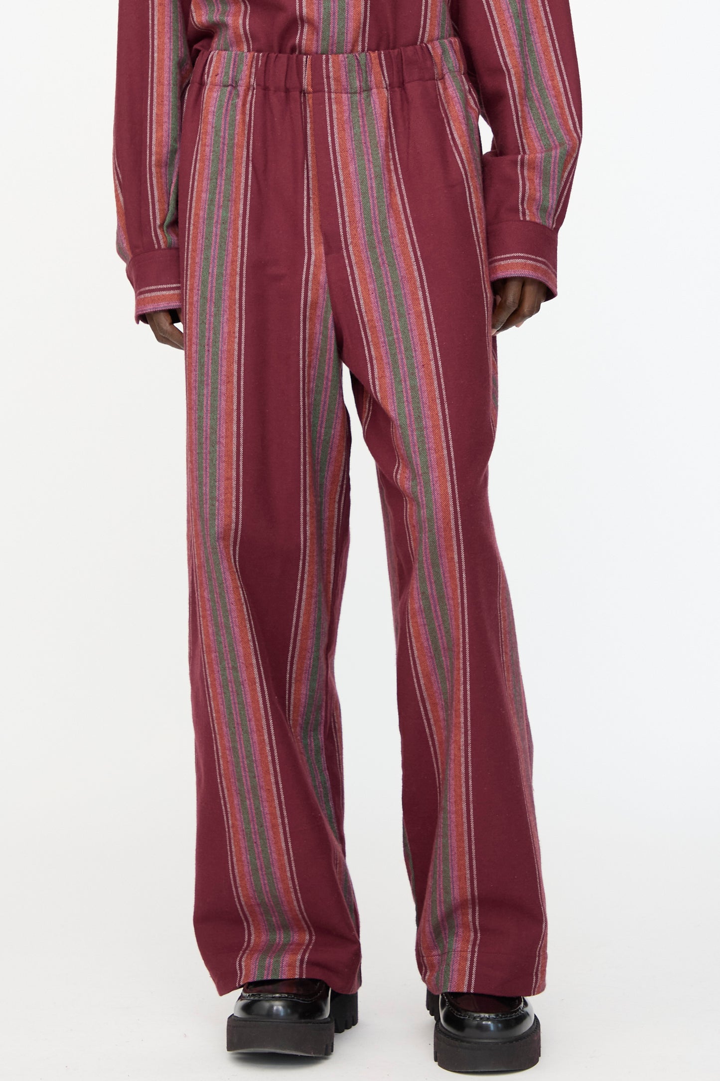 A person wears Baserange's Claude Pants in Maroon Stripe—loose-fitting, wide-leg pull-on pants with vertical multicolored stripes—paired with a matching long-sleeve top and black platform shoes against a plain background.