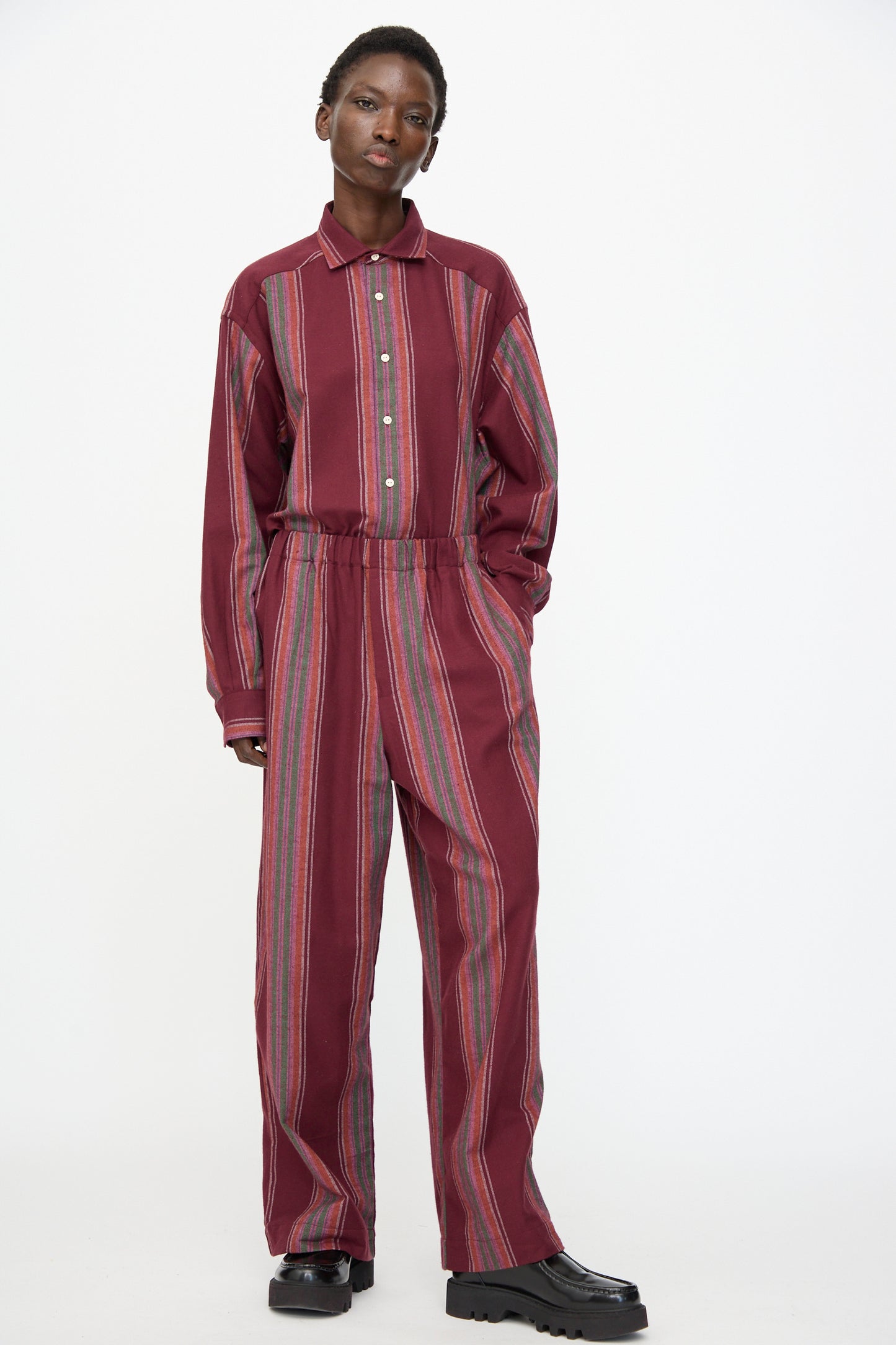 A person stands against a white background wearing Baserange's Claude Pants in Maroon Stripe with black shoes.