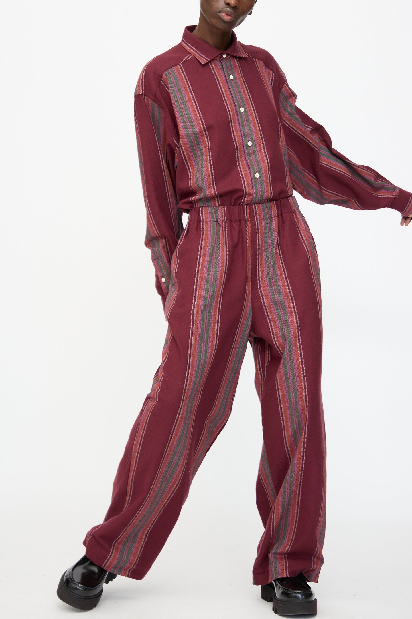 Model wears Baserange Claude Pants in Maroon Stripe—wide-leg, pull-on style with vertical multicolor stripes—paired with a loose, long-sleeve maroon shirt, standing against a plain white background.