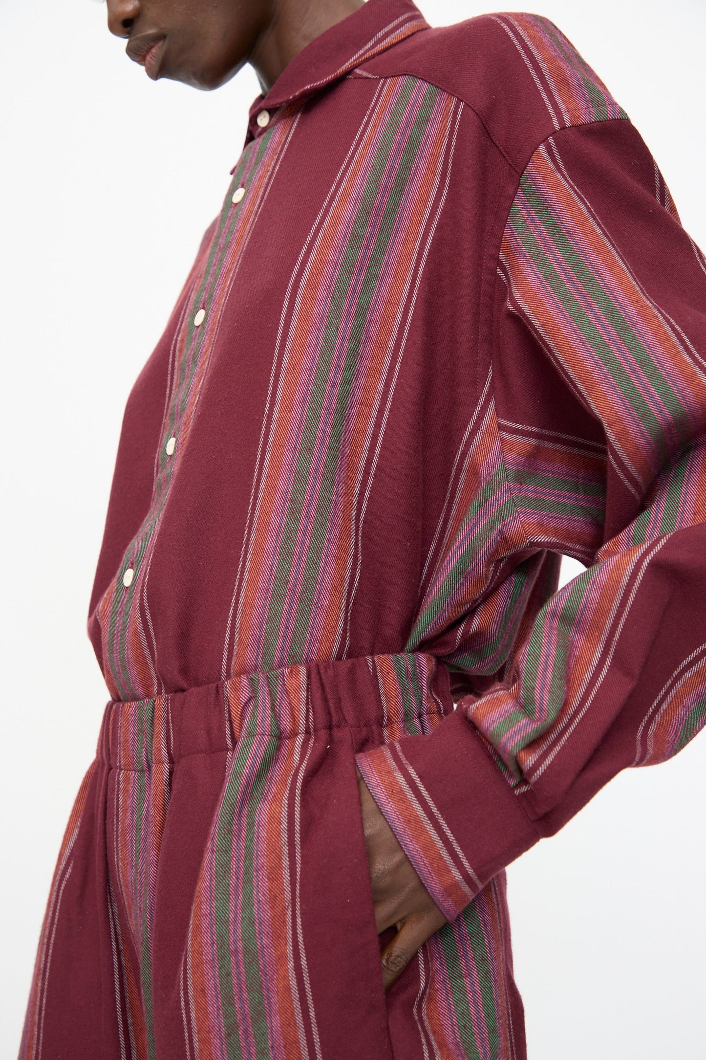Model wears Baserange Claude Pants in Maroon Stripe—wide leg, organic cotton, vertical multicolored stripes, and white buttons—shown shoulders to mid-thigh with hand in pocket.