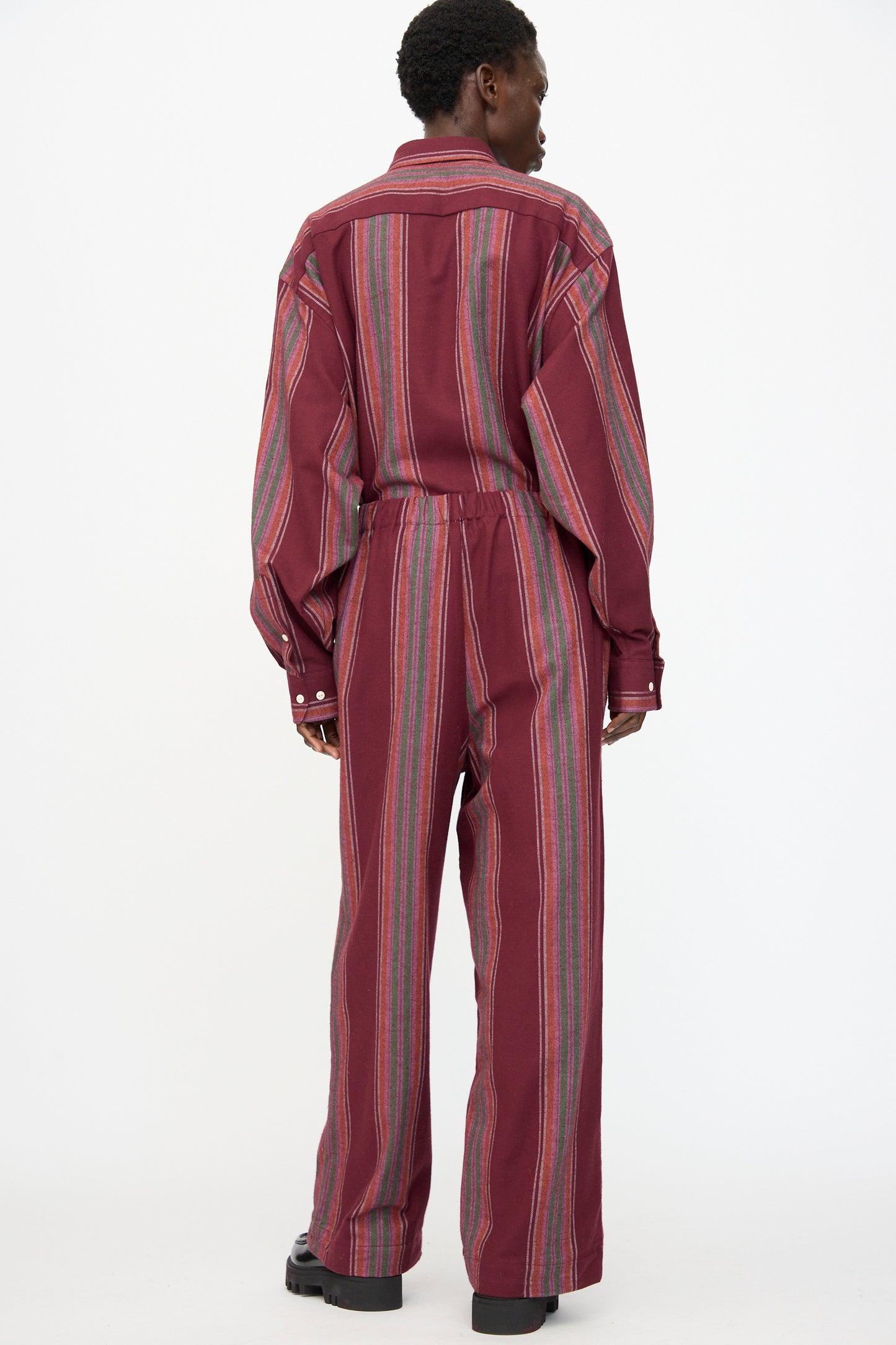 A person stands facing away in Baserange Claude Pants in Maroon Stripe—loose-fitting, wide-leg, organic cotton pants with vertical multicolored stripes and long sleeves—against a plain white background.