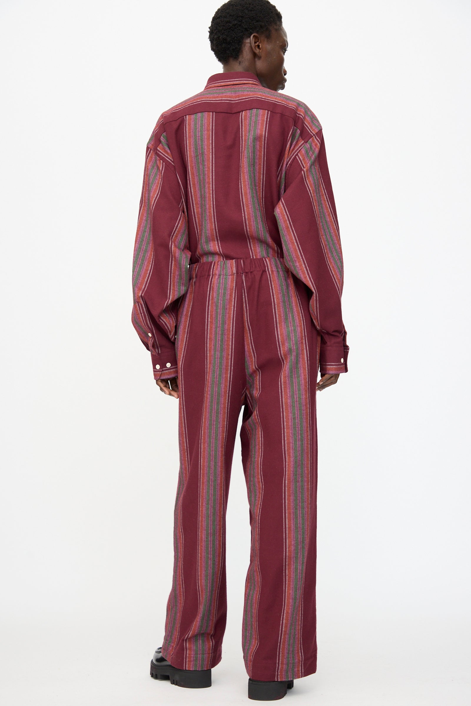 A person stands facing away in Baserange Claude Pants in Maroon Stripe—loose-fitting, wide-leg, organic cotton pants with vertical multicolored stripes and long sleeves—against a plain white background.