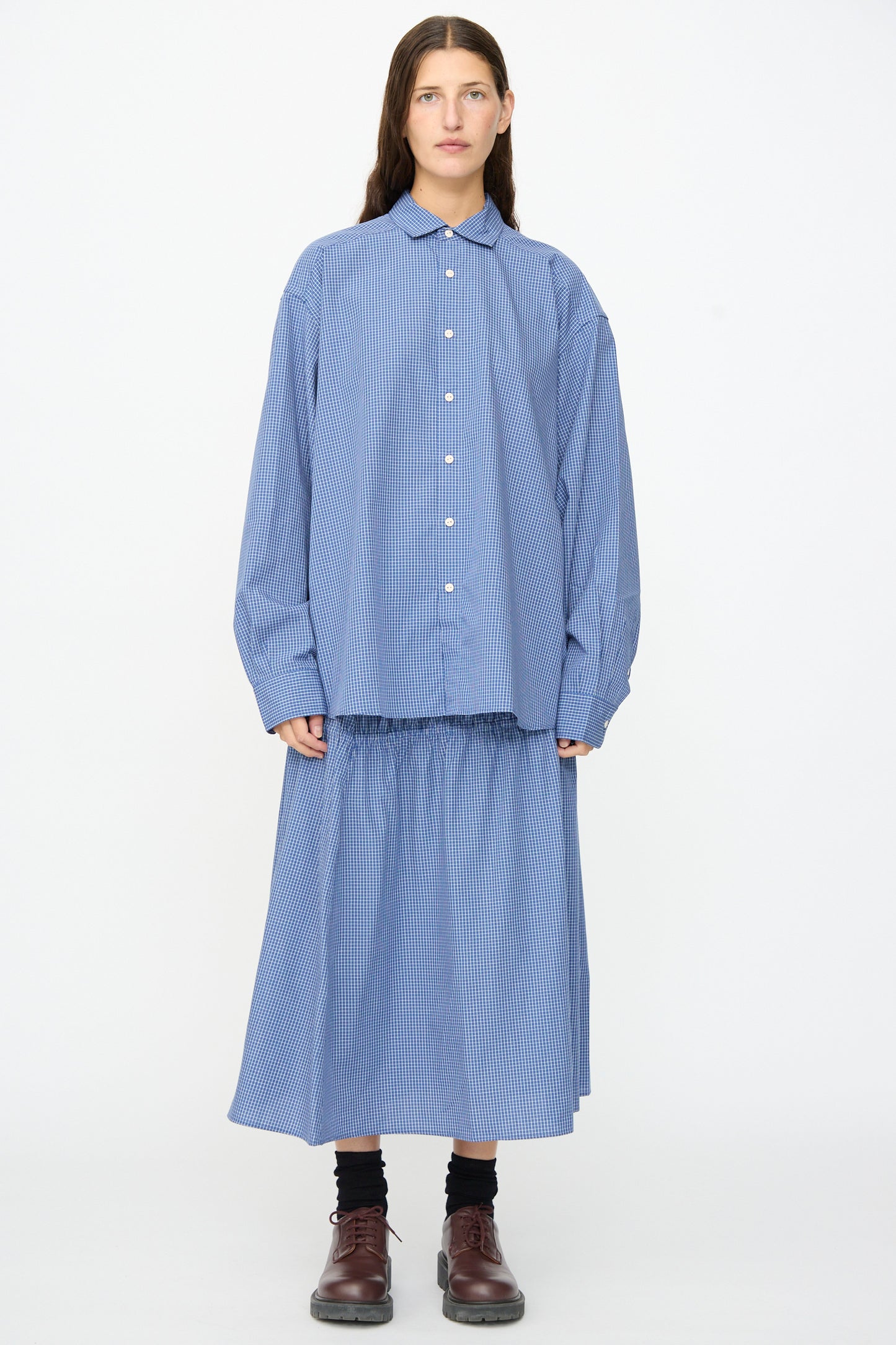 A person stands against a white background wearing the Baserange Claude Shirt in Ace Check—a relaxed fit blue striped cotton shirt—paired with a matching skirt made in Portugal, black socks, and brown shoes.