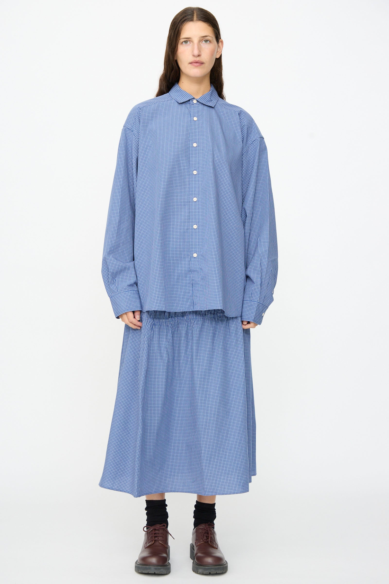 A person stands against a white background wearing the Baserange Claude Shirt in Ace Check—a relaxed fit blue striped cotton shirt—paired with a matching skirt made in Portugal, black socks, and brown shoes.