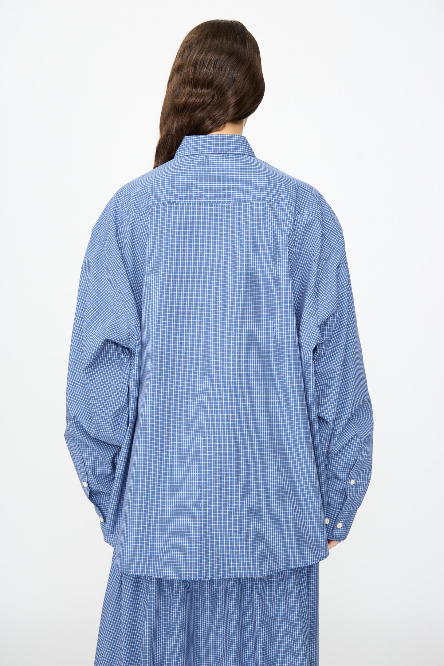 A person wearing the Baserange Claude Shirt in Ace Check—a relaxed fit, blue and white checkered button-up with long sleeves, made in Portugal—is photographed from behind against a plain white background.