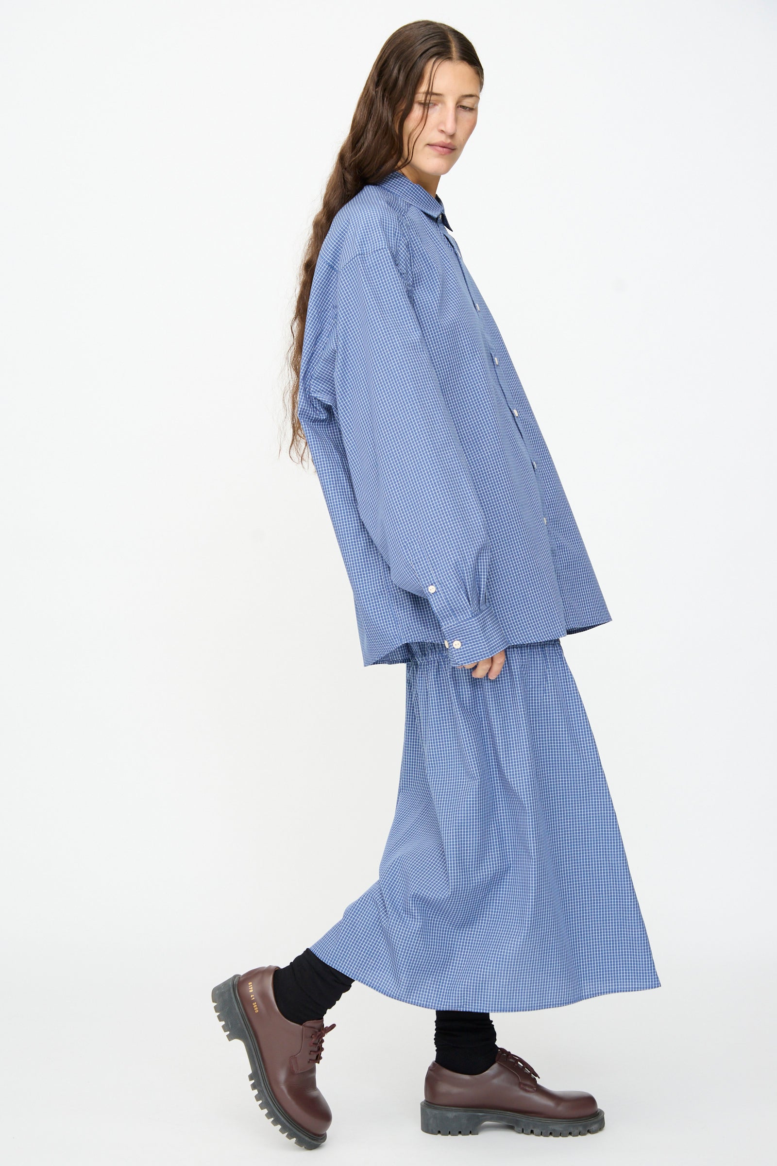 A person wearing the Baserange Claude Shirt in Ace Check—a relaxed fit, blue pinstripe shirt made in Portugal—paired with a matching skirt, black socks, and brown platform shoes, standing against a plain white background.