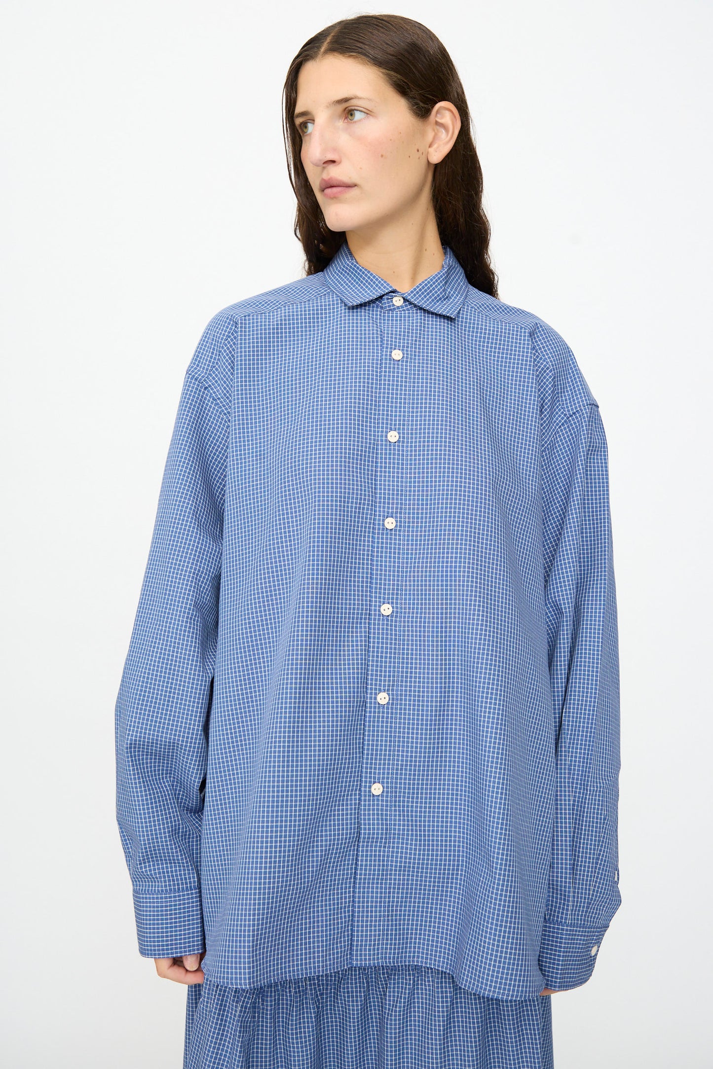 A person wearing the Baserange Claude Shirt in Ace Check, an oversized blue checkered button-up with a relaxed fit, stands against a plain white background and looks slightly to the side.