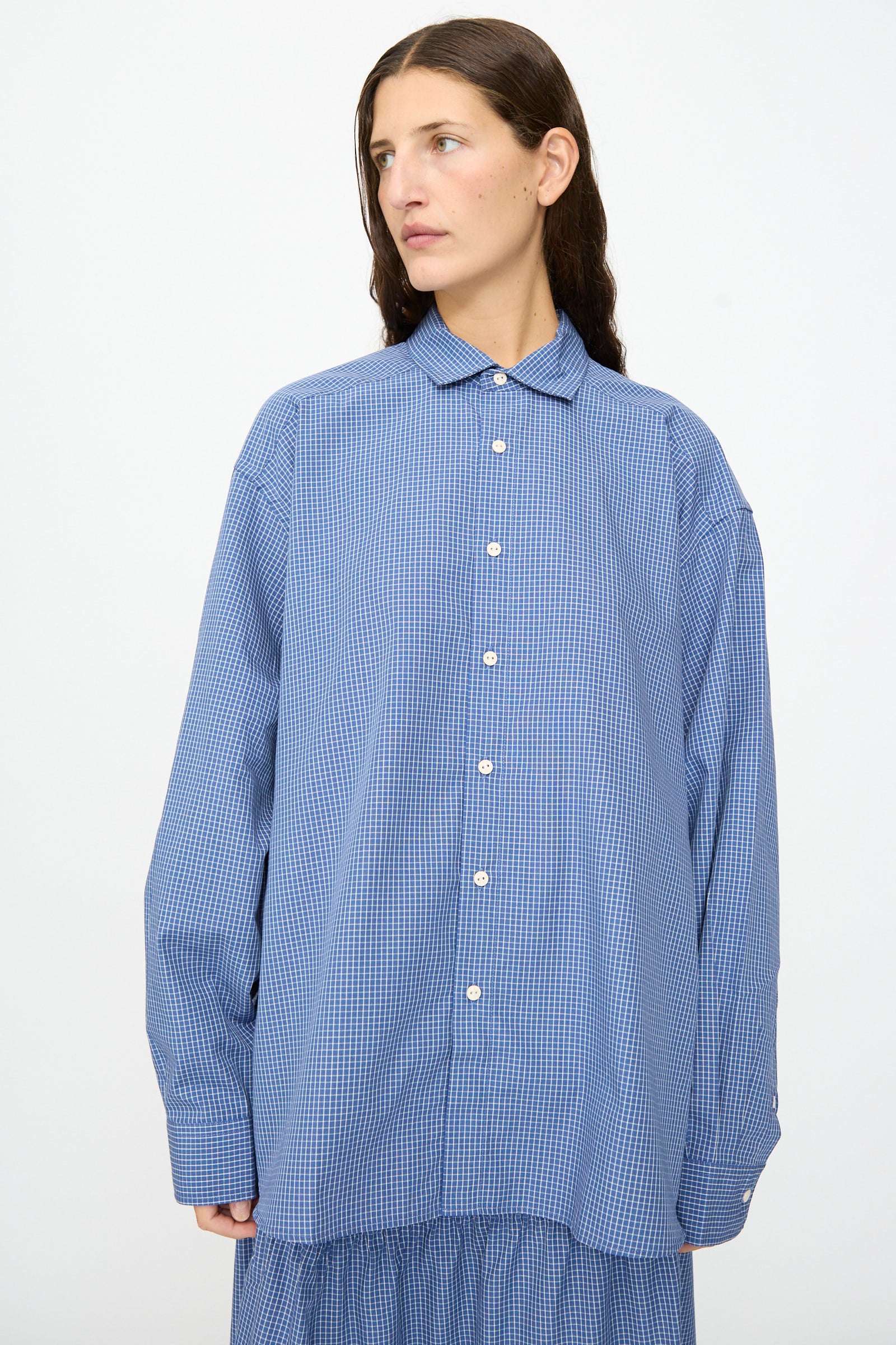 A person wearing the Baserange Claude Shirt in Ace Check, an oversized blue checkered button-up with a relaxed fit, stands against a plain white background and looks slightly to the side.