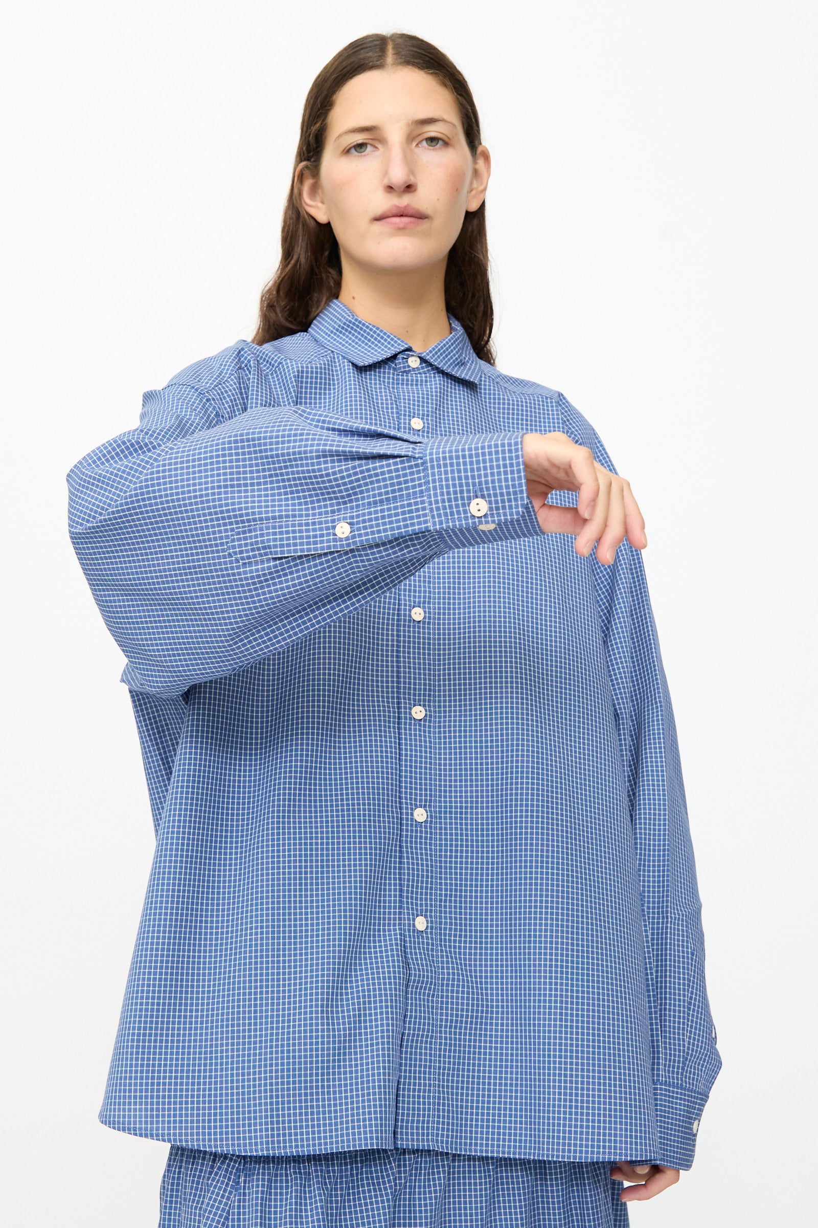 A person wears the Baserange Claude Shirt in Ace Check—an oversized, relaxed fit blue checkered button-up—paired with matching bottoms, standing against a plain white background with one arm raised and bent.