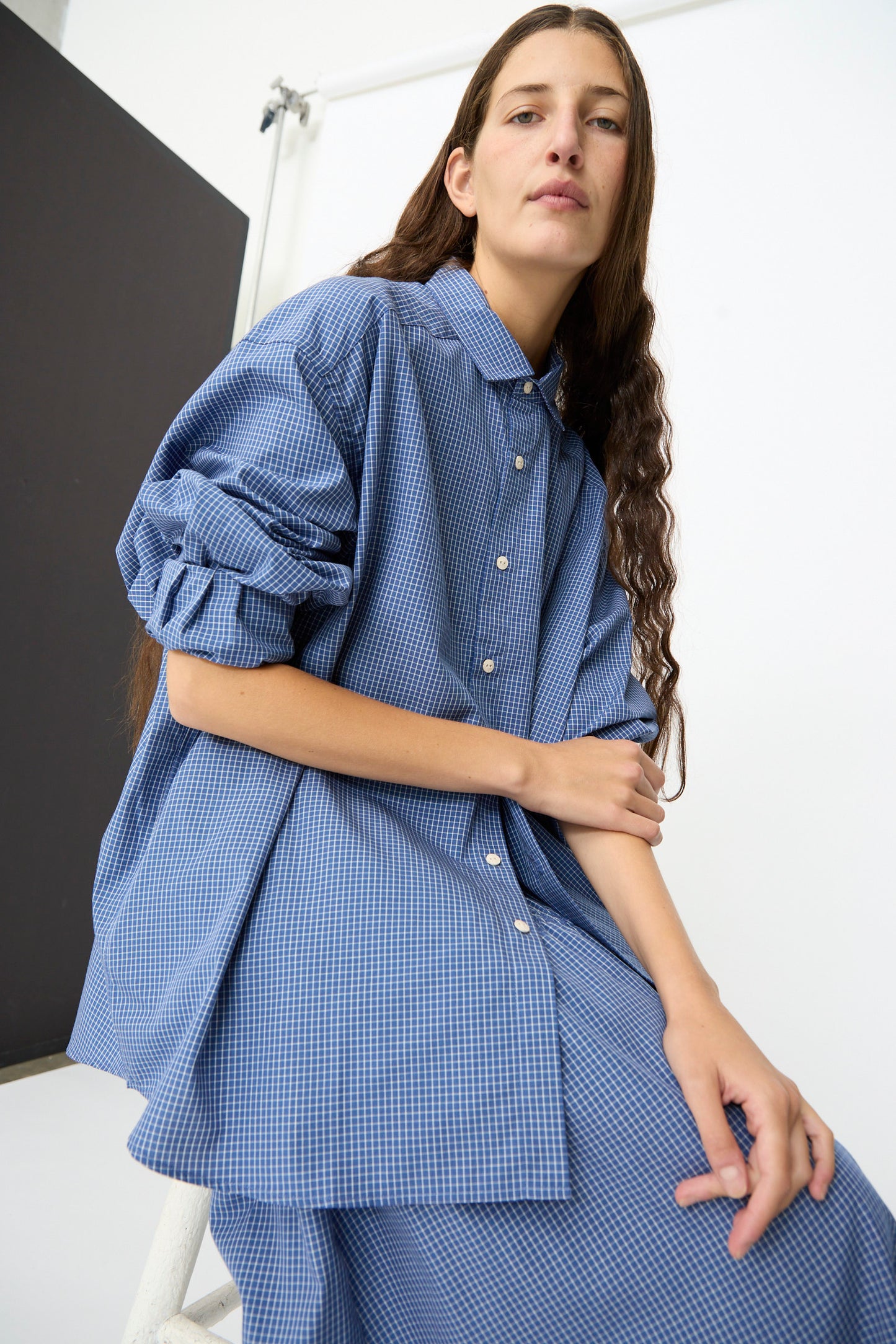 A person with long brown hair wears the Baserange Claude Shirt in Ace Check, an oversized blue checkered shirt with a relaxed fit, paired with a matching skirt, while sitting on a white stool against a plain background.