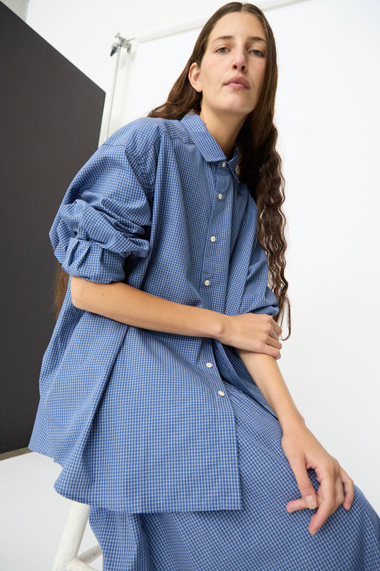 A person with long brown hair wears the Baserange Claude Shirt in Ace Check, an oversized blue checkered shirt with a relaxed fit, paired with a matching skirt, while sitting on a white stool against a plain background.