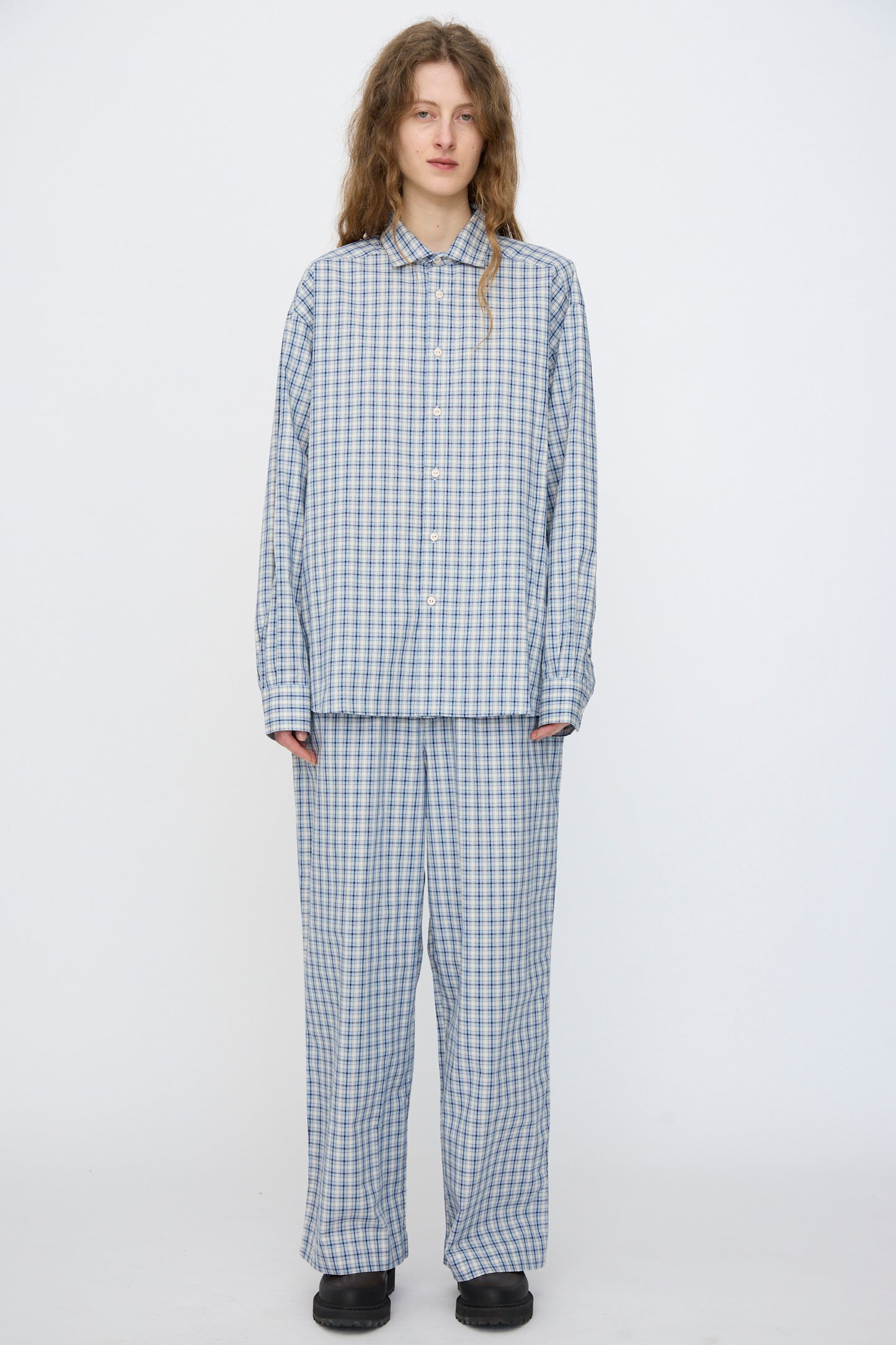 A person stands against a plain background, wearing Baserange's Claude Shirt in Blue Check made from organic cotton, paired with matching pants and black shoes.