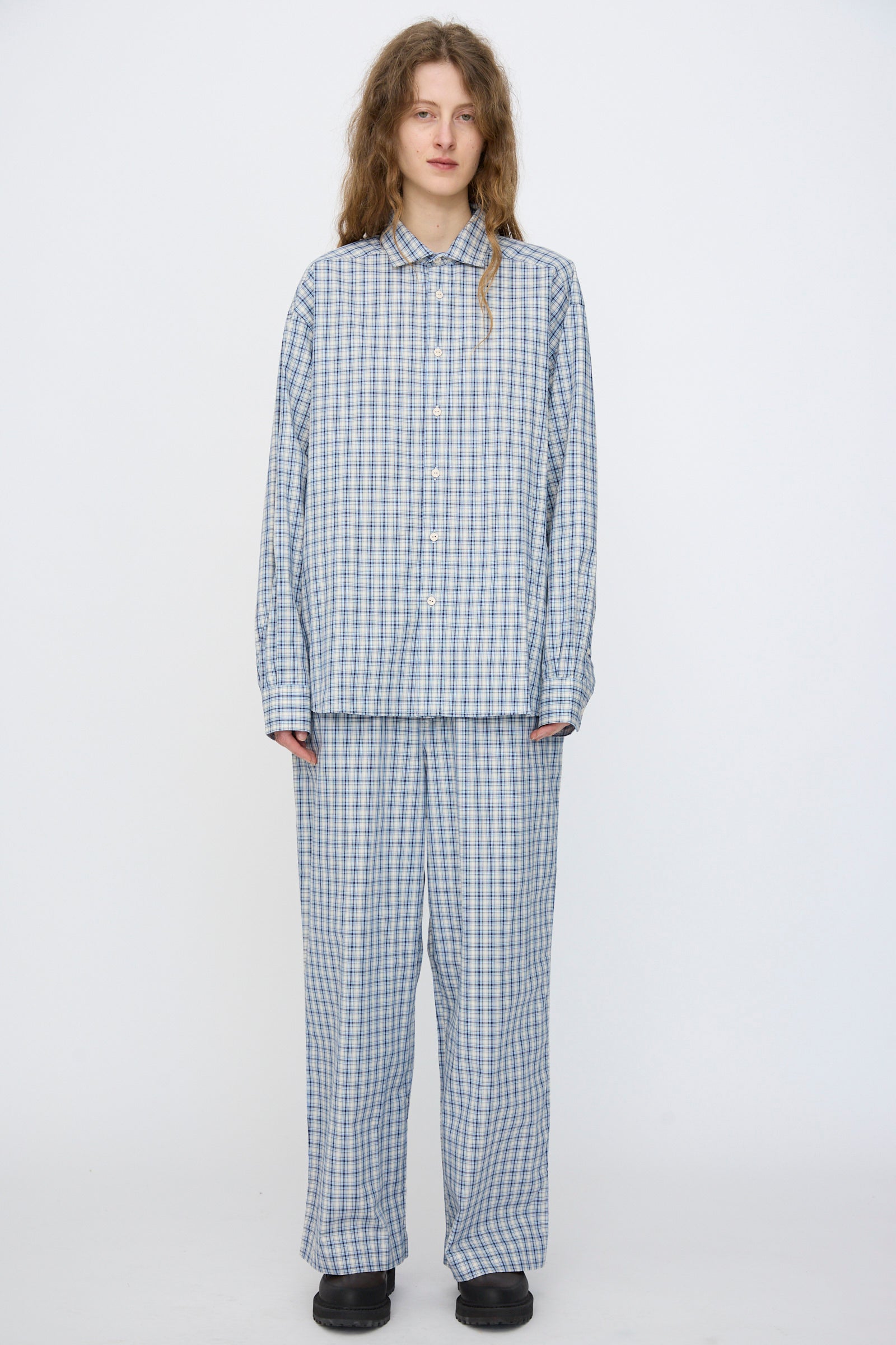 A person stands against a plain background, wearing Baserange's Claude Shirt in Blue Check made from organic cotton, paired with matching pants and black shoes.