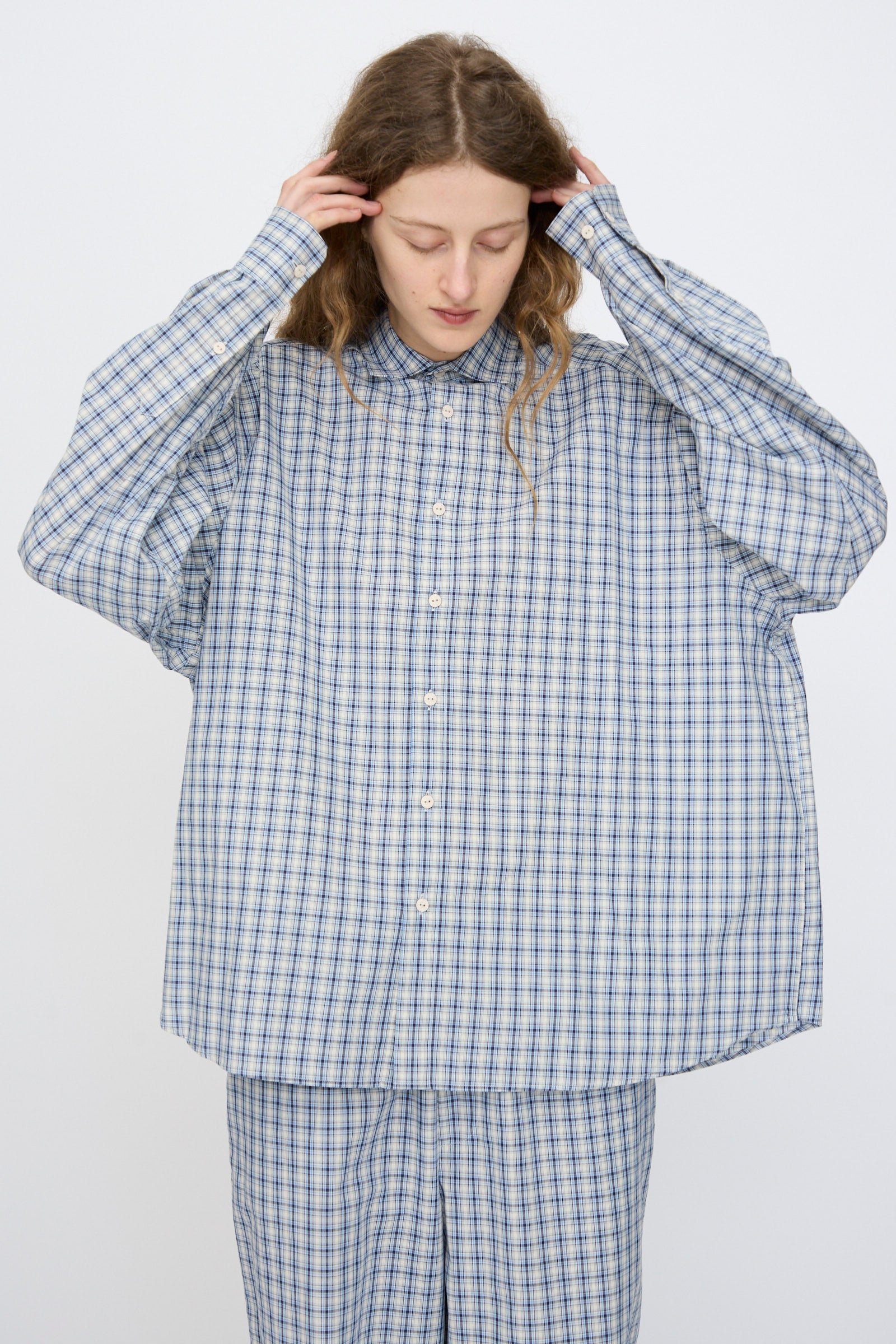 A person wearing the Baserange Claude Shirt in Blue Check stands against a plain background, looking down and touching their head.