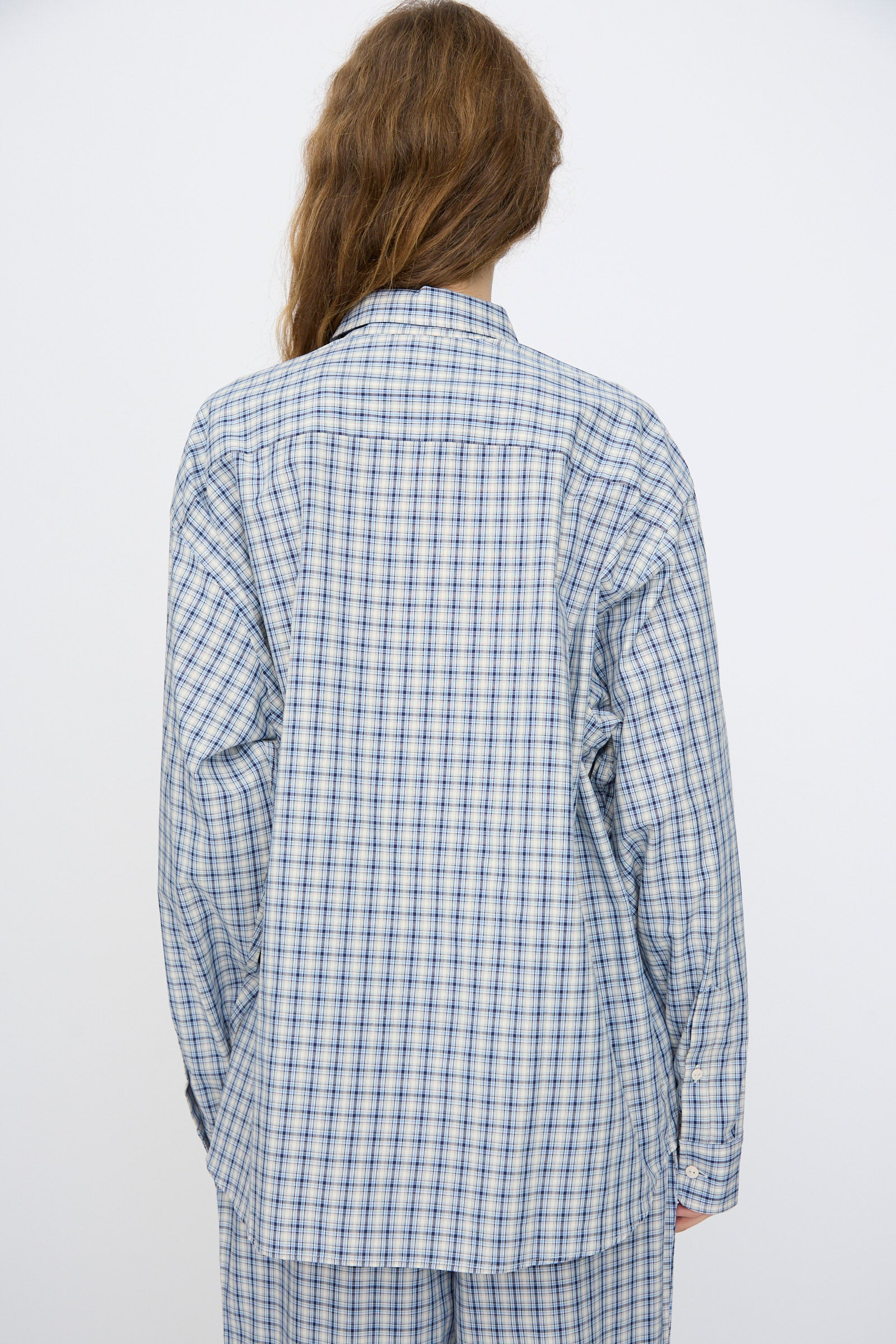 A person with long brown hair faces away, wearing the Baserange Claude Shirt in Blue Check with matching pants, set against a plain light background.